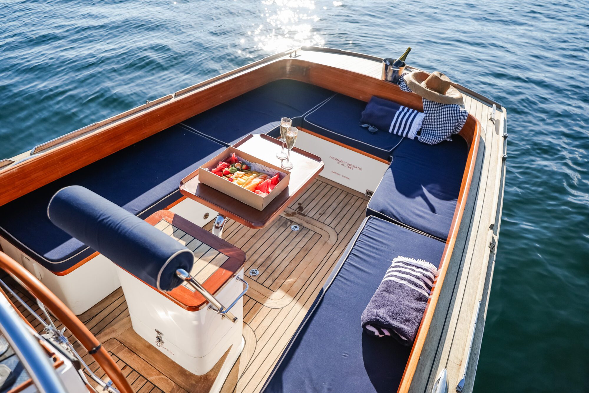 A luxurious boat deck with blue cushioned seating, a tray of fruit, a glass of champagne, and a bottle in an ice bucket. Sunlight sparkles on calm water, and a hat and blanket rest on the seats.