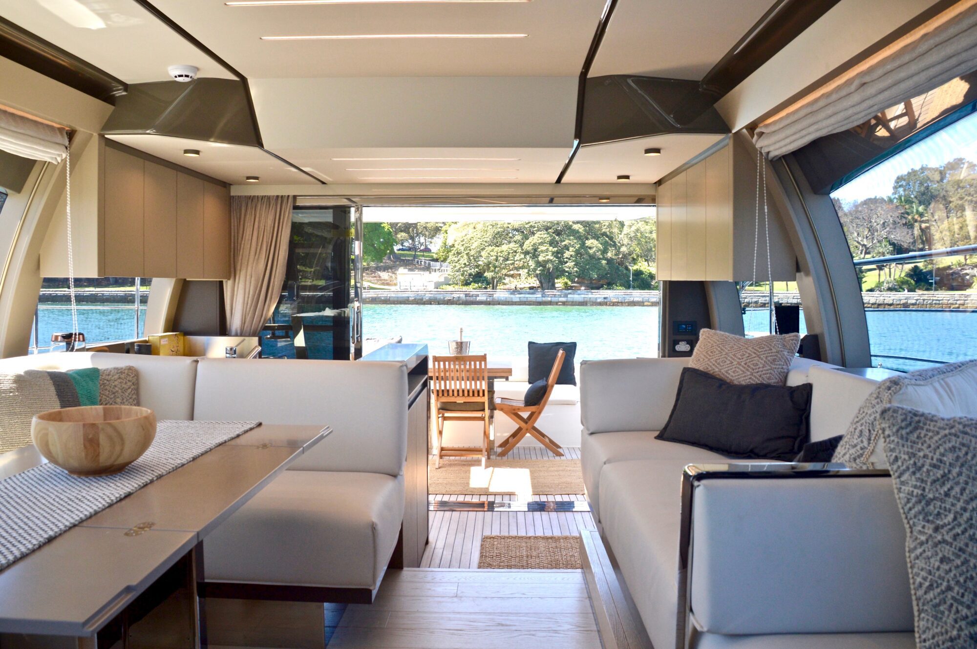 Interior of a modern yacht with light-colored sofas, a dining table, and large windows. The view outside shows a wooden deck, chairs, water, and trees under a sunny sky.