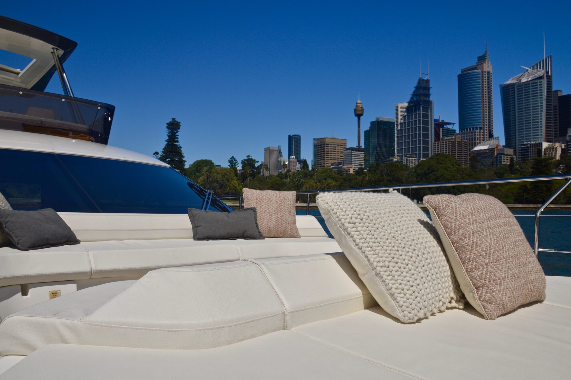 View from a yacht’s sun deck with cushioned seating and pillows, overlooking a city skyline with tall buildings and trees under a clear blue sky.