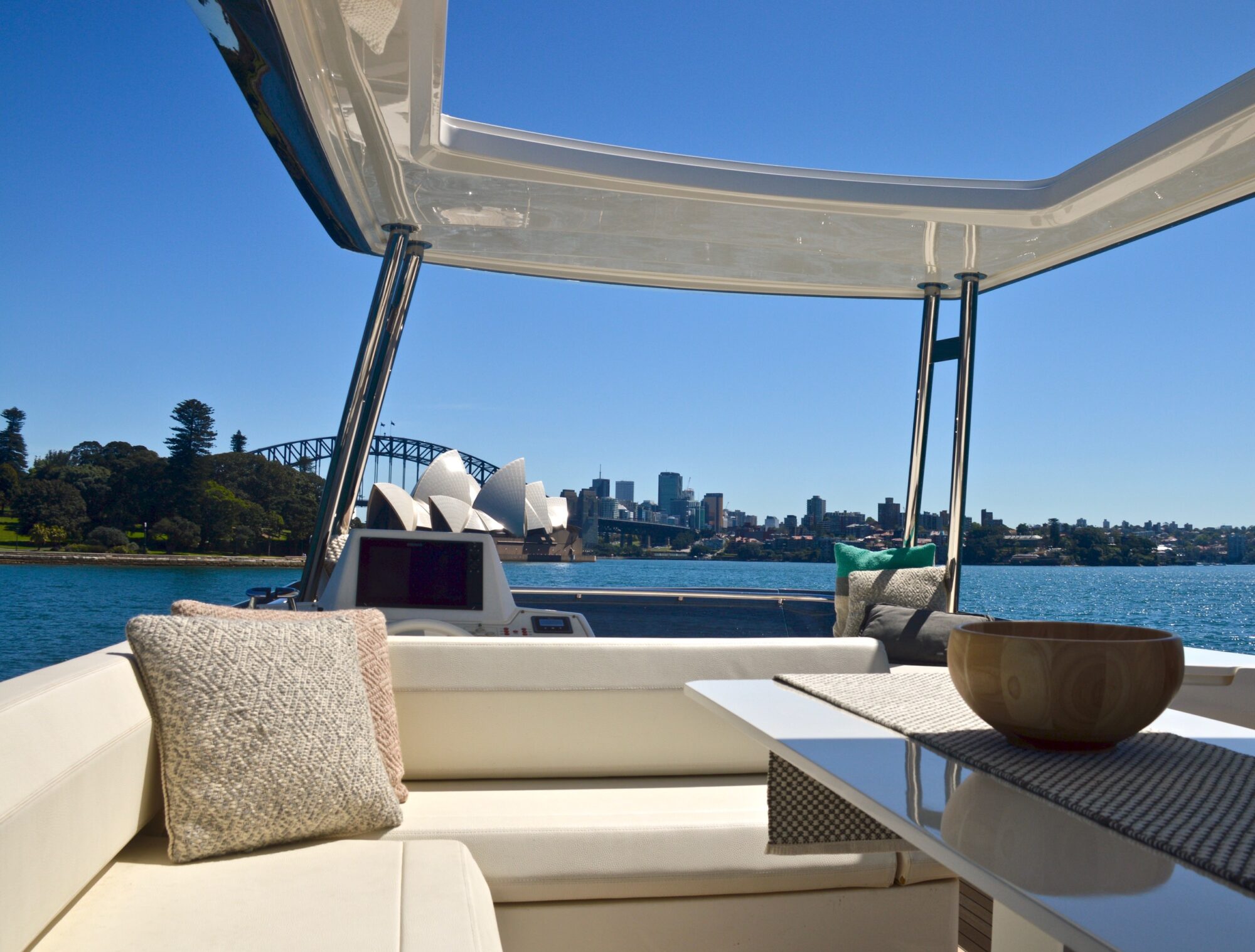 A view from a yacht’s shaded seating area, looking out over Sydney Harbour with the Sydney Opera House and Harbour Bridge visible against a clear blue sky. Cushions and a bowl rest on the yacht’s white seats and table.