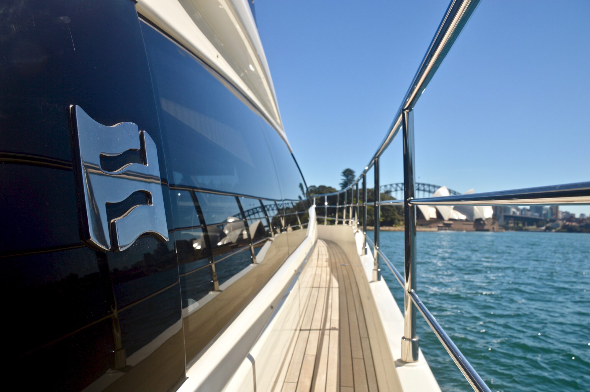 View from the deck of a yacht on blue water, with a city skyline, Sydney Opera House, and Sydney Harbour Bridge visible in the background under a clear blue sky.