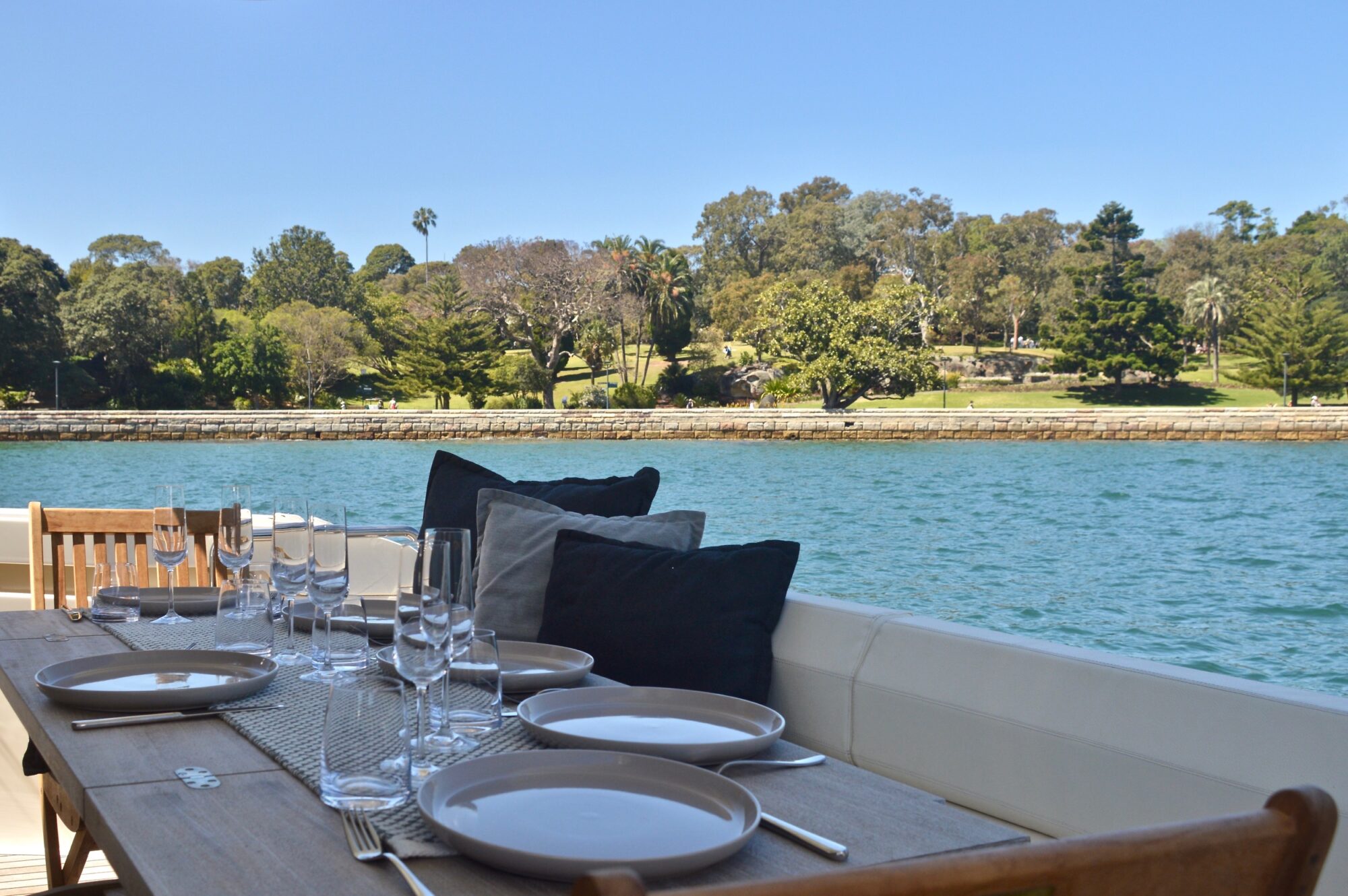 A dining table set with plates, glasses, and cushions is on a boat overlooking calm blue water and a green, tree-lined shoreline under a clear sky.