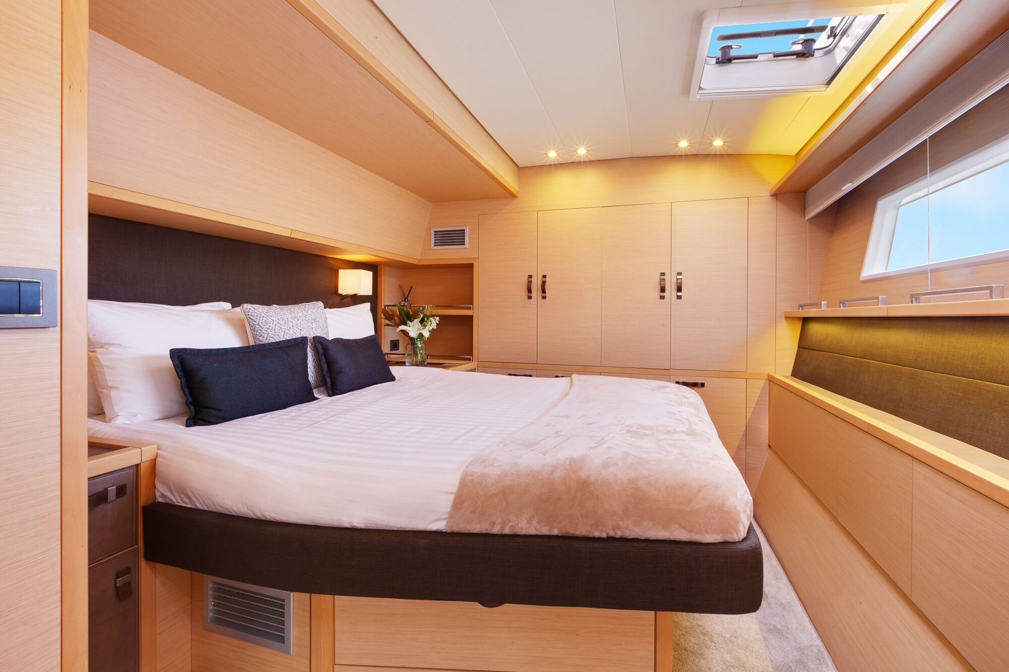 A modern yacht bedroom with light wood paneling, a large neatly made bed with white linens and black accent pillows, built-in cabinets, and a skylight letting in natural light.