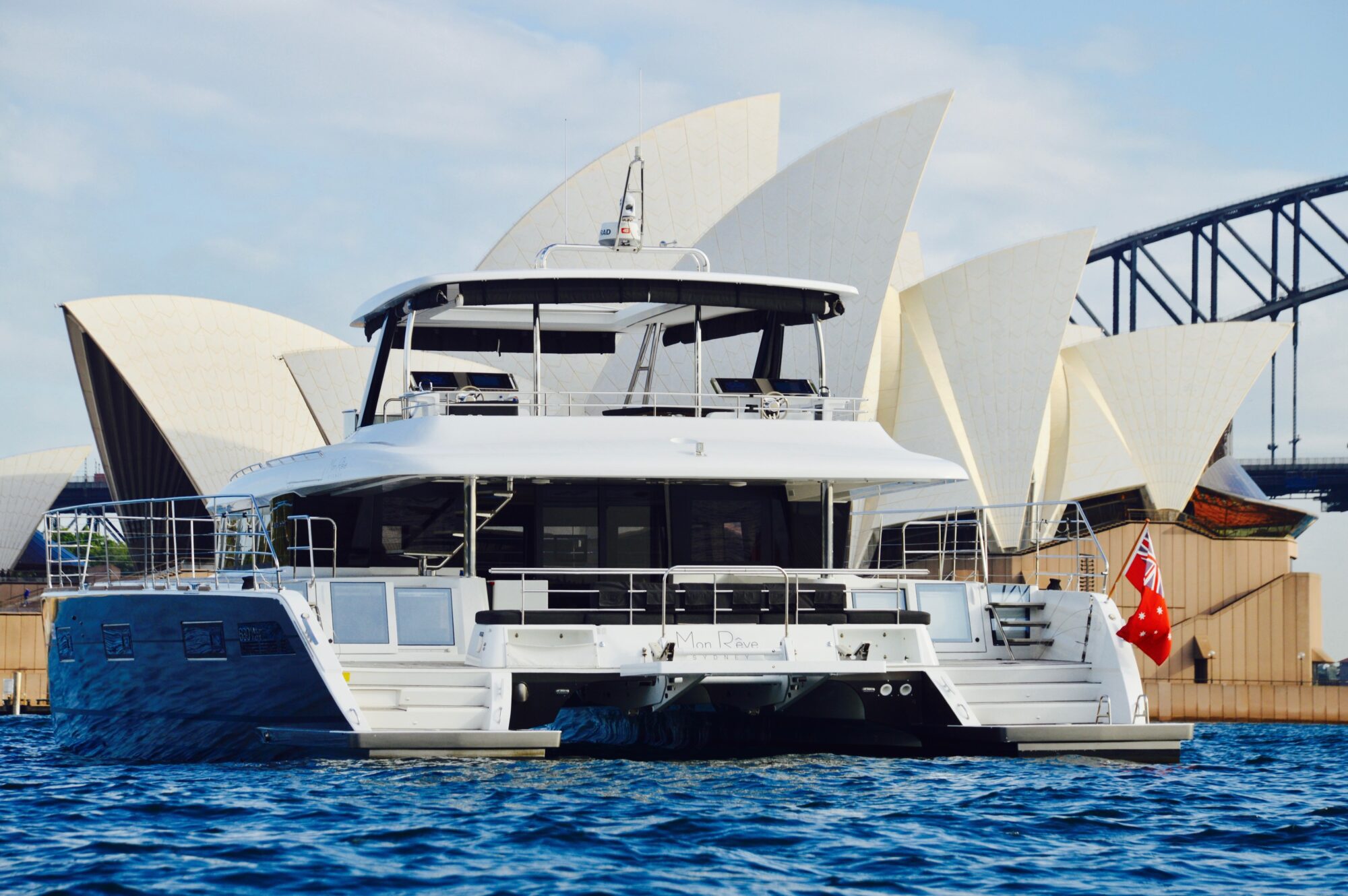 A modern white catamaran yacht floats on blue water, with the Sydney Opera House and part of Sydney Harbour Bridge visible in the background under a partly cloudy sky.