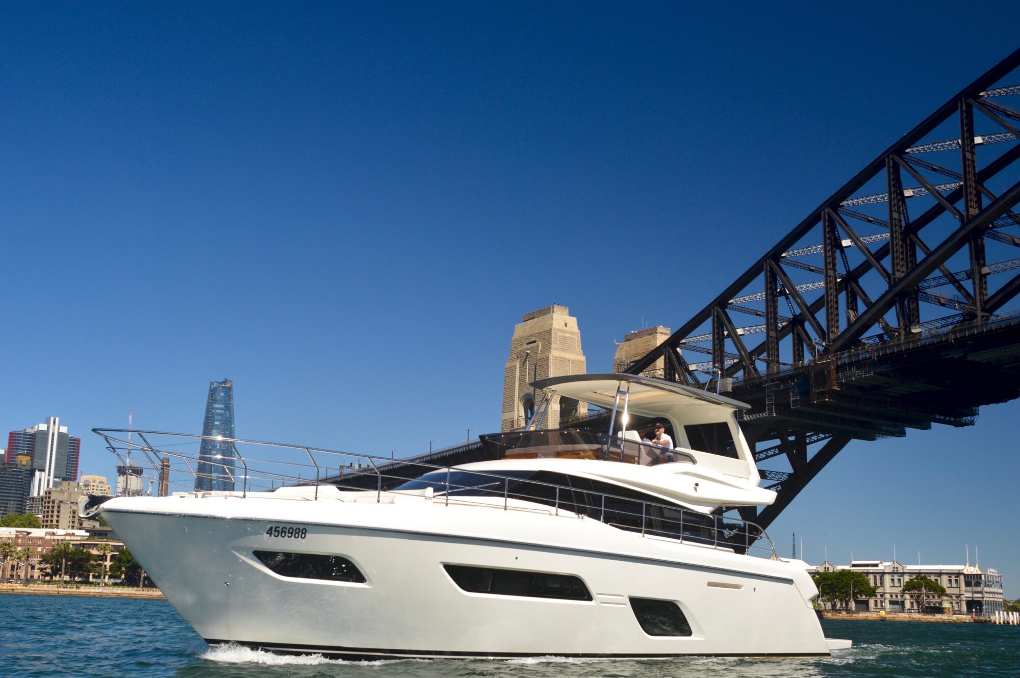 A white yacht sails near the Sydney Harbour Bridge on a sunny day, with modern city buildings visible in the background against a clear blue sky.