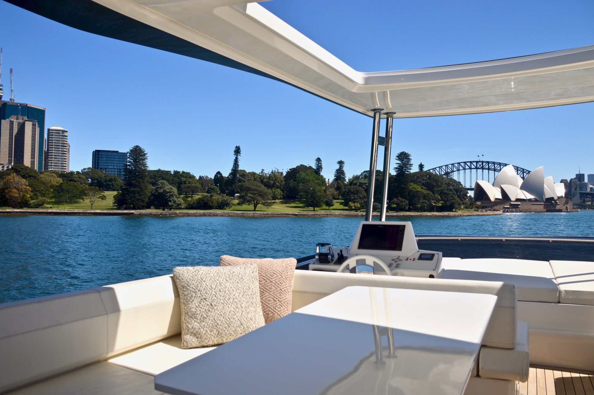 View from a yacht’s outdoor seating area with cushions and a table, overlooking Sydney Harbour, the Sydney Opera House, Harbour Bridge, and waterfront greenery under a clear blue sky.