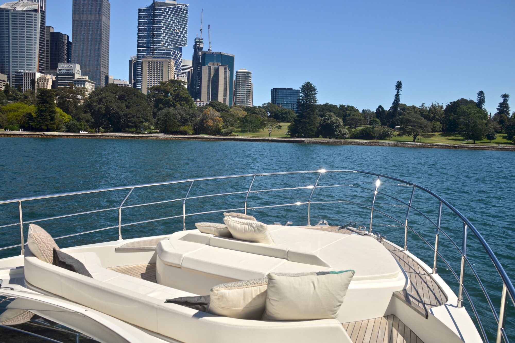 A luxurious yacht with white cushioned seating is cruising on blue water near a city skyline with tall buildings and trees on a sunny day.