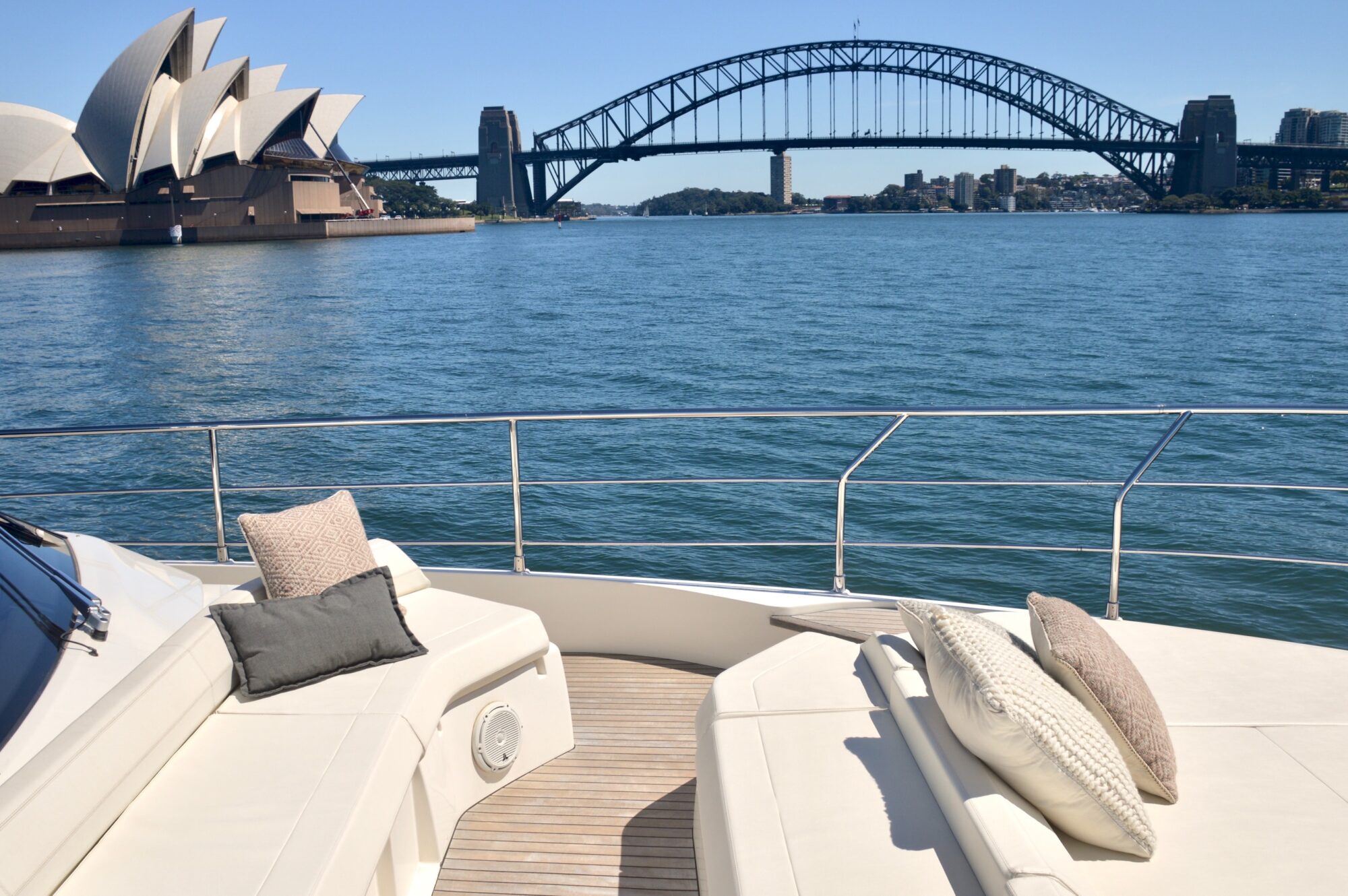 View from a yacht’s deck with cushioned seating, overlooking Sydney Opera House and Sydney Harbour Bridge on a sunny day. Calm water and clear blue sky in the background.