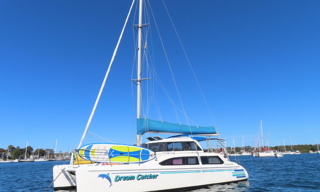 A white catamaran sailboat named "Dream Catcher" floats on calm blue water under a clear sky, with a paddleboard secured on top and other boats visible in the background.