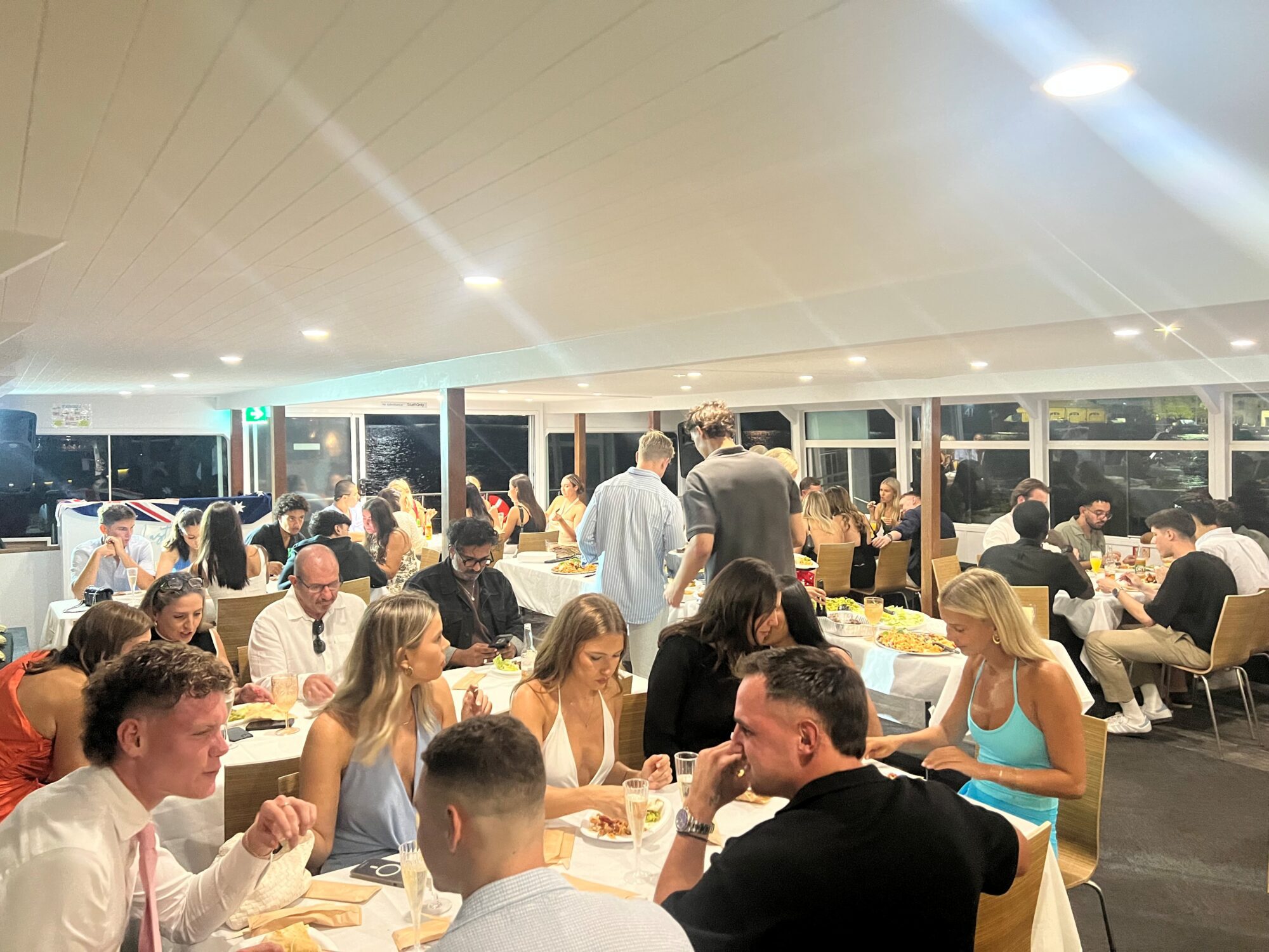 A large group of people dressed in semi-formal attire sit at tables in a brightly lit restaurant, eating, drinking, and socializing during a lively dinner event.