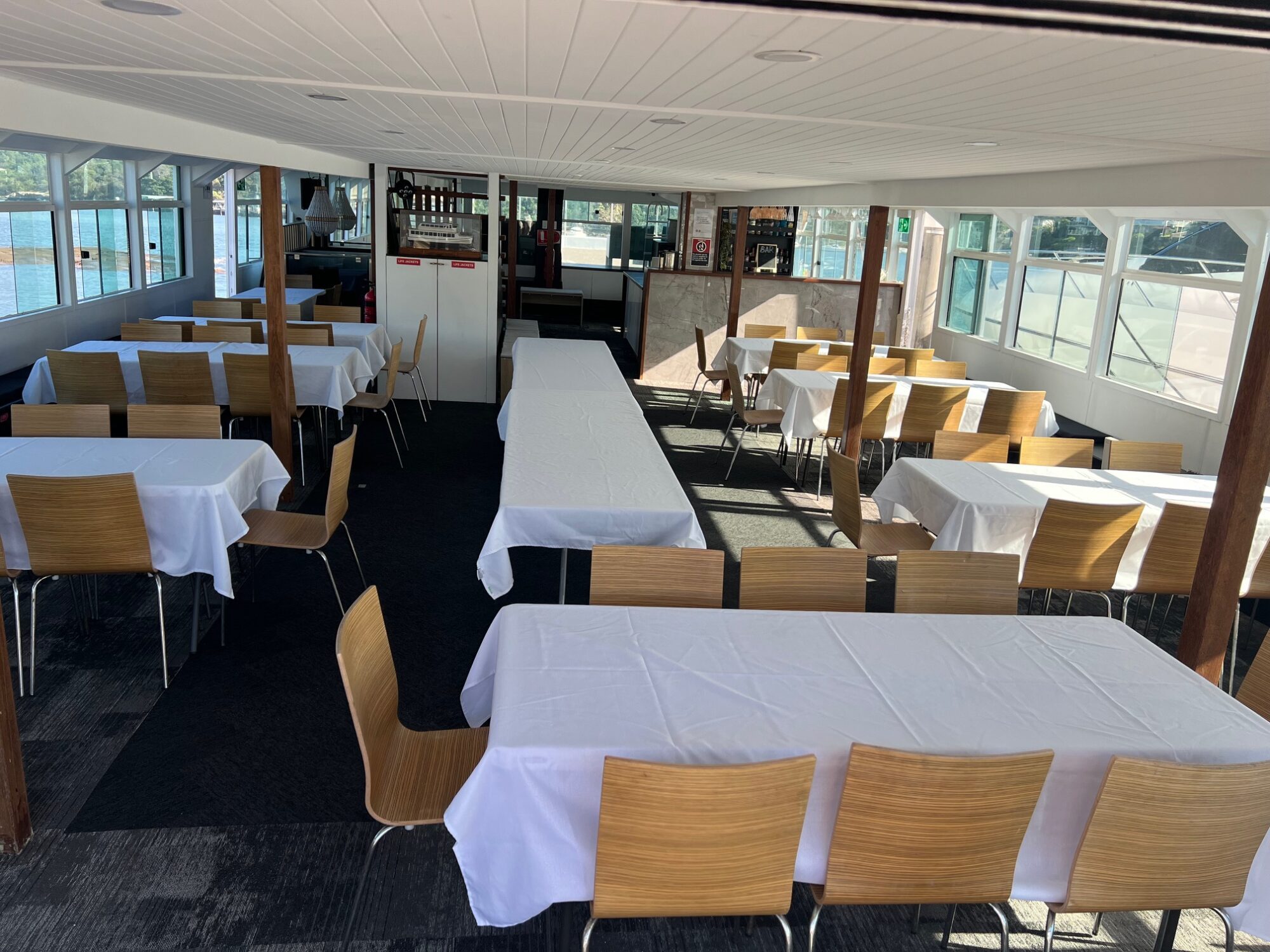 Interior of a boat with large windows, set up for dining. Several rectangular tables covered with white tablecloths are surrounded by wooden chairs, with sunlight streaming through the windows.