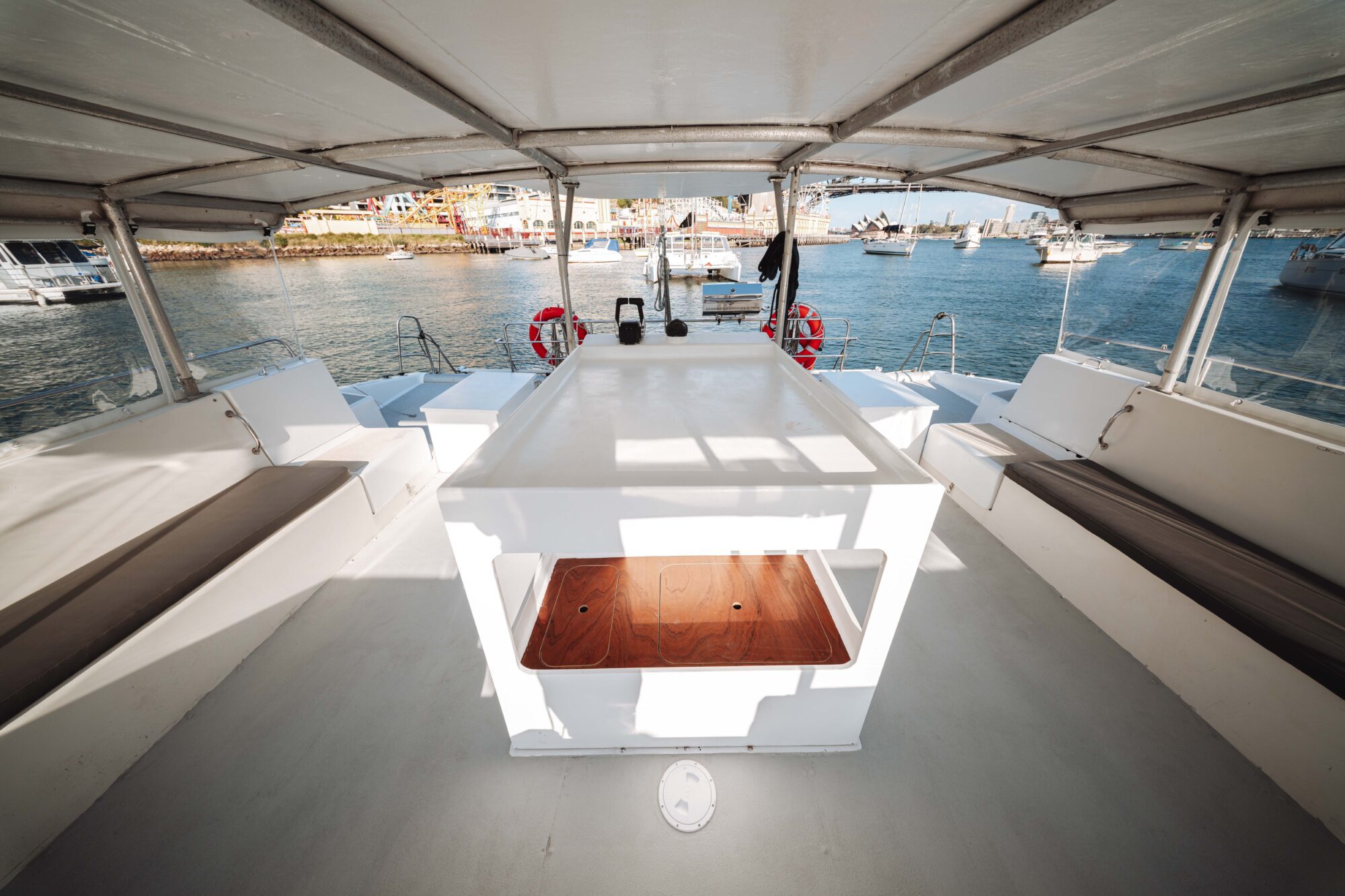 View from the shaded deck of a boat, featuring white benches on both sides, a central table, and a marina filled with boats in the background under a bright sky.