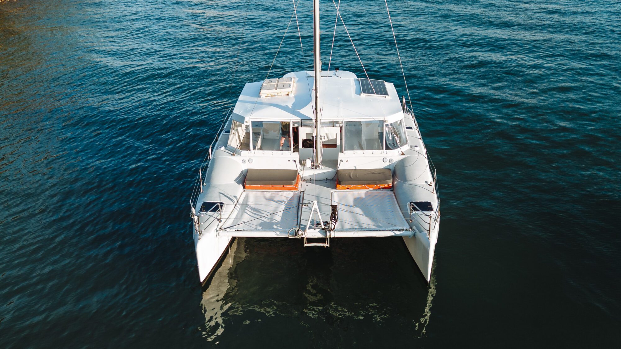 A white catamaran boat with two hulls floats on calm blue water, seen from above. The deck has seating areas and solar panels on the roof. The boat is anchored and there are no people visible.