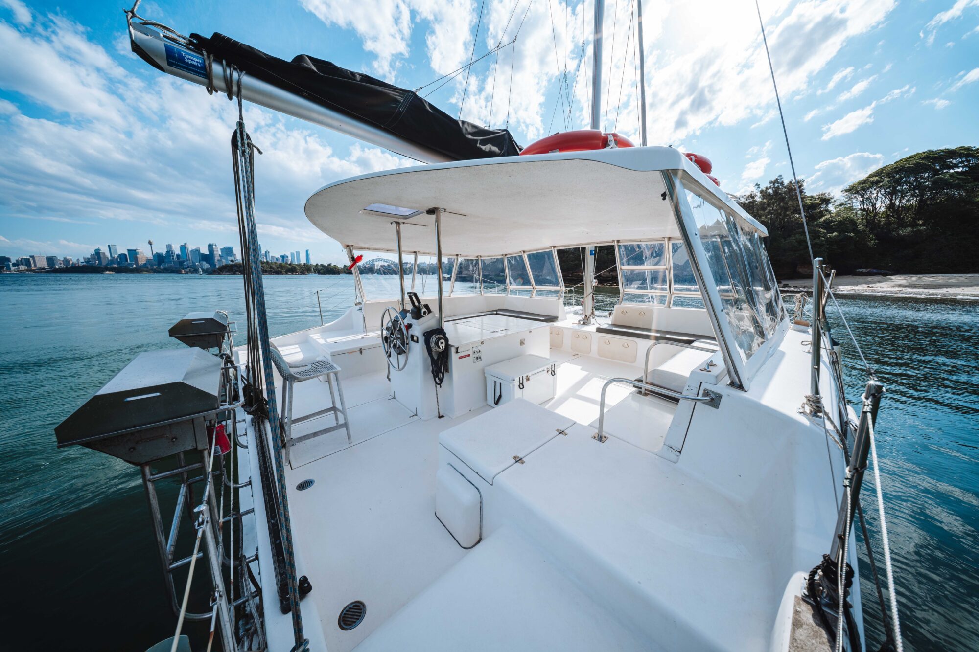A spacious, white yacht deck with seating, steering wheel, and clear canopy, floating on calm water near a shoreline, with a city skyline visible in the background under a partly cloudy sky.