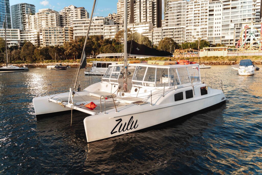 A white catamaran named "Zulu" floats on calm water near a city shoreline, with high-rise buildings and several other boats in the background on a sunny day.