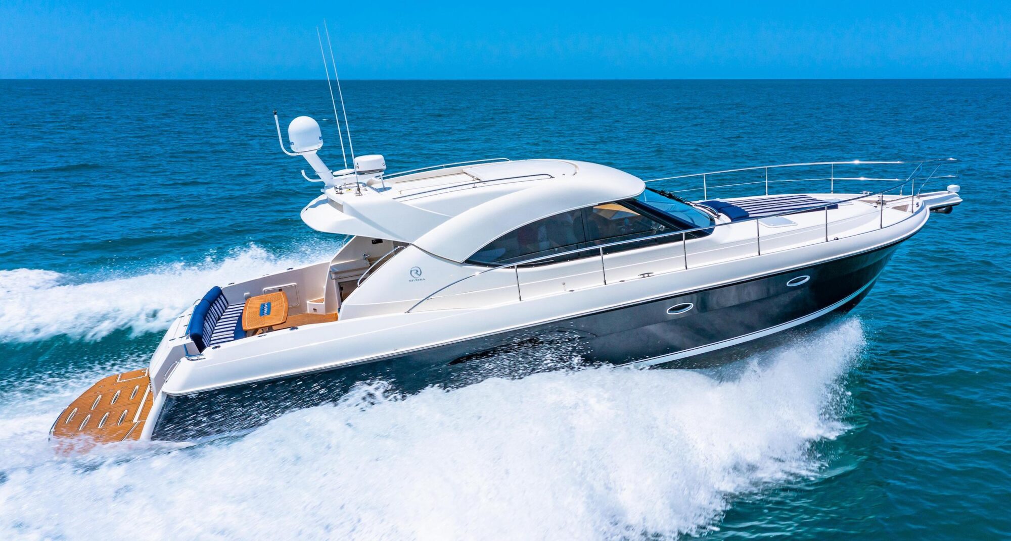 A sleek white and black motor yacht speeds across a calm blue ocean, creating foamy waves behind it under a clear sky.