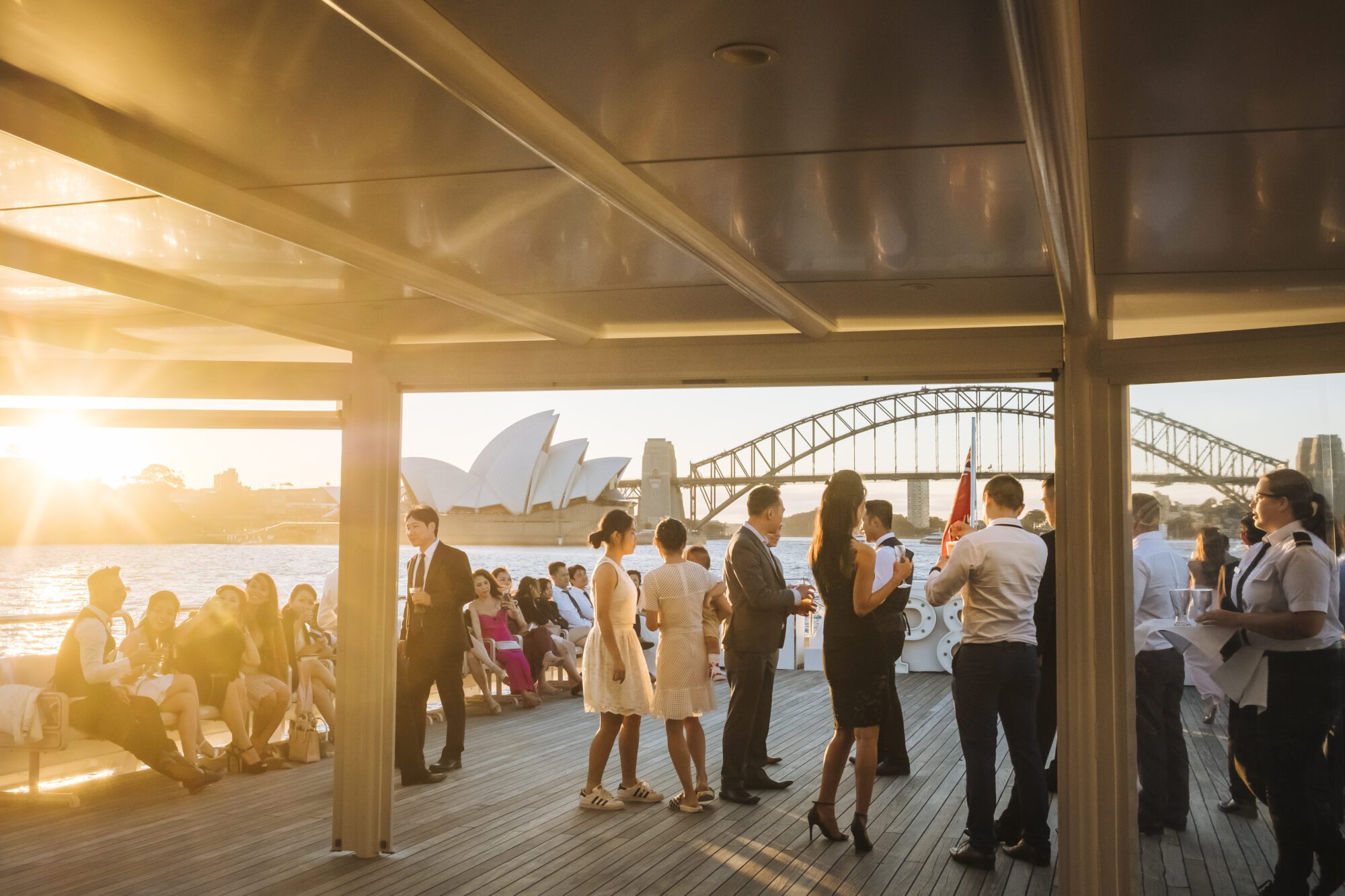 People socialize on a boat deck at sunset, with the Sydney Opera House and Harbour Bridge visible in the background. The scene is lively, with guests dressed in semi-formal attire, enjoying drinks and conversation.