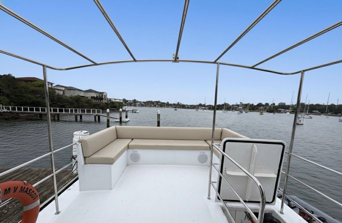 View from a boat’s upper deck with cushioned seating, a safety railing, and a life ring. The boat overlooks a calm harbor with several anchored boats and houses along the shoreline under a clear blue sky.