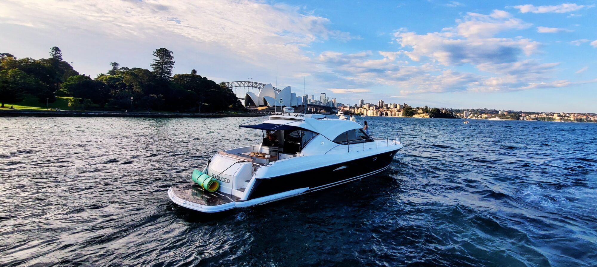 A white and navy yacht sails on a harbor with city skyline, green trees, and the Sydney Opera House and Harbour Bridge visible in the background under a partly cloudy blue sky.