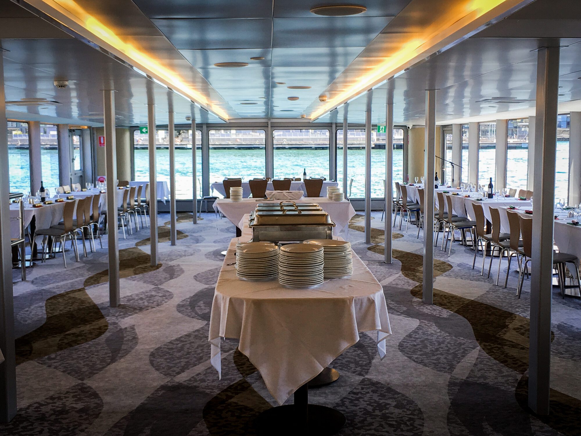A spacious, elegant dining area on a boat, set with white tablecloths and neatly arranged chairs, overlooks large windows revealing water outside. Plates and serving dishes are prepared on tables in the center aisle.