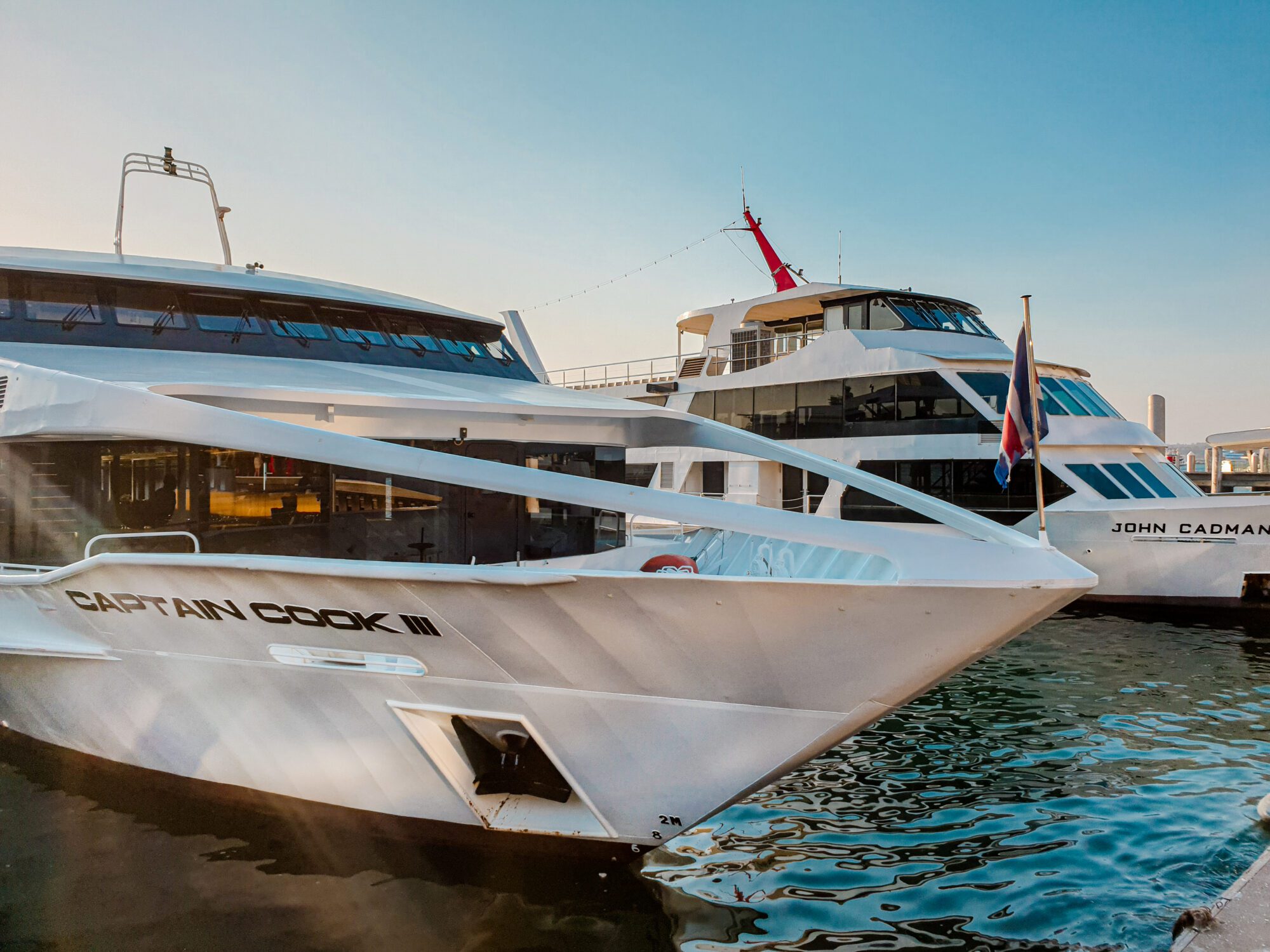 Two modern white cruise boats, "CAPTAIN COOK III" and "JOHN CADMAN," are docked side by side in a harbor under a clear blue sky. The water reflects their hulls.