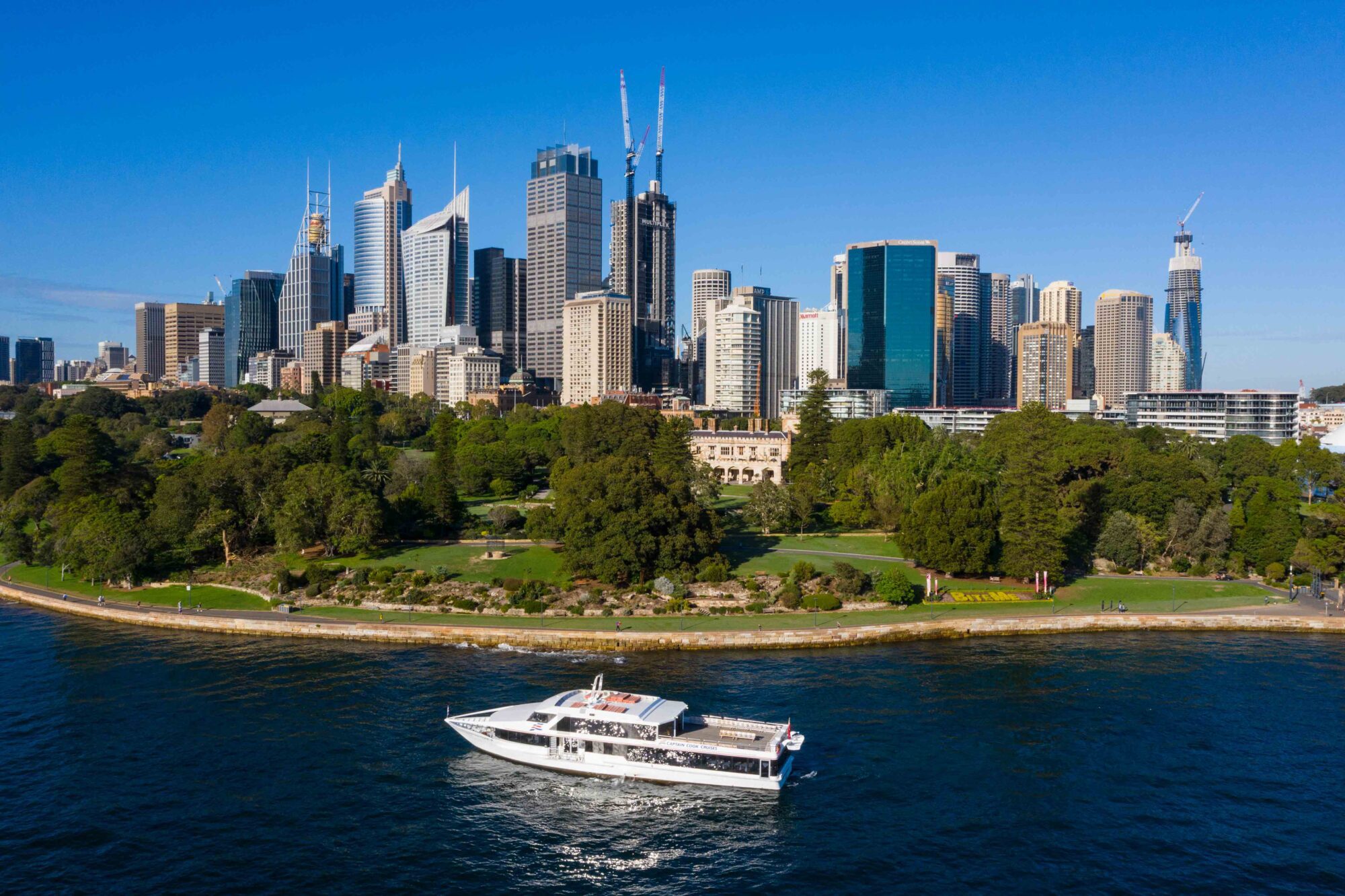 A white yacht sails along the water in front of a lush green park, with a city skyline of tall modern skyscrapers under a clear blue sky in the background.