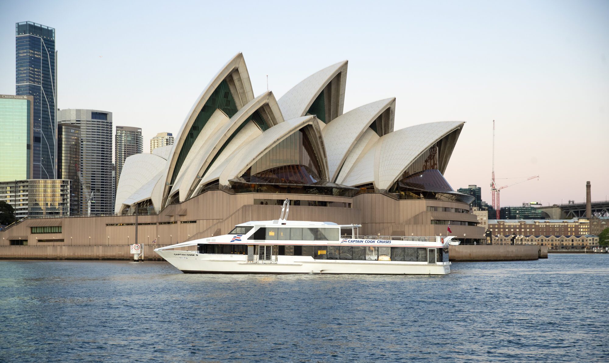A white ferry sails on the water in front of the Sydney Opera House, with its iconic sail-shaped roof, and modern city buildings in the background at sunset.