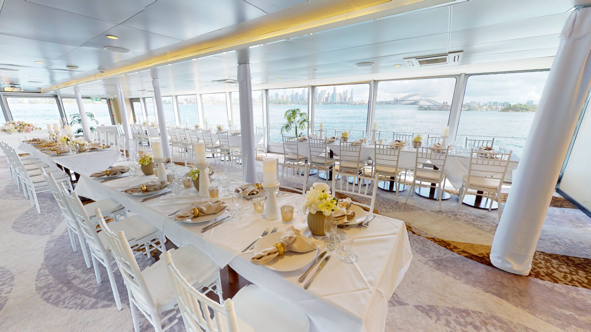 Elegant dining area on a boat with large windows overlooking water and a distant city skyline. Tables are set with white linens, flowers, candles, and napkins folded on plates. White chairs surround the tables.