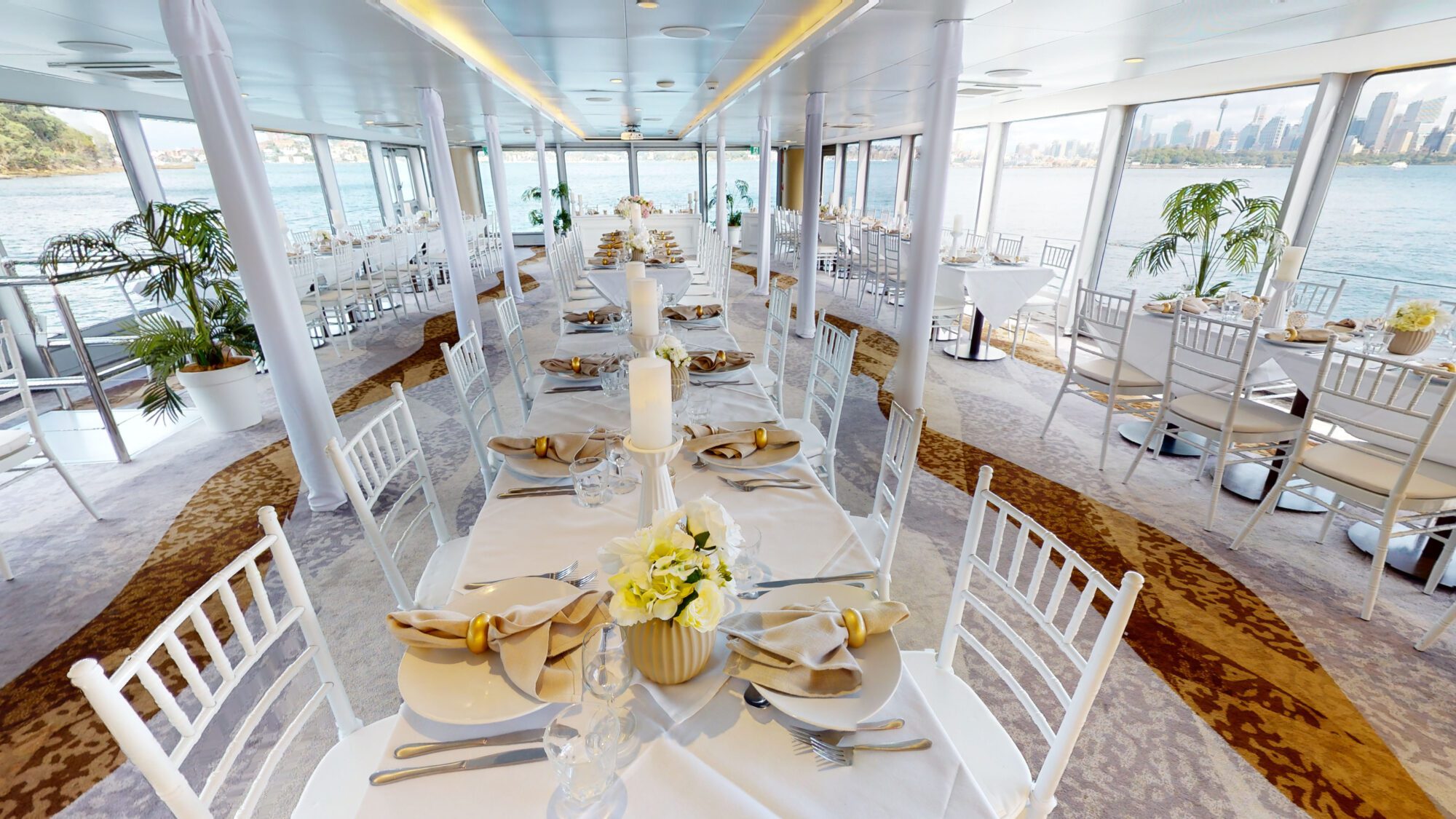 Elegant dining area on a boat with white tables and chairs, gold napkins, floral centerpieces, large windows, and views of water and city skyline in the background.