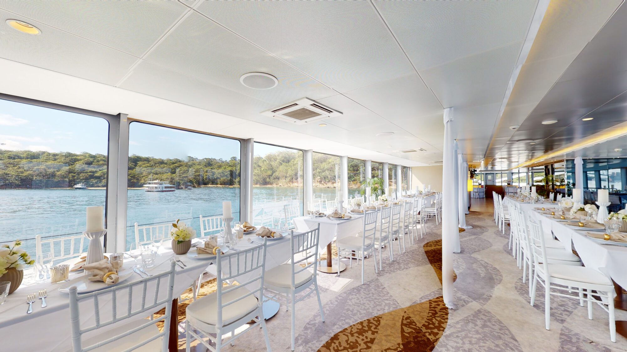 A bright, elegant dining area on a boat features white tables and chairs set for a meal, with large windows offering scenic views of calm water and green trees outside.