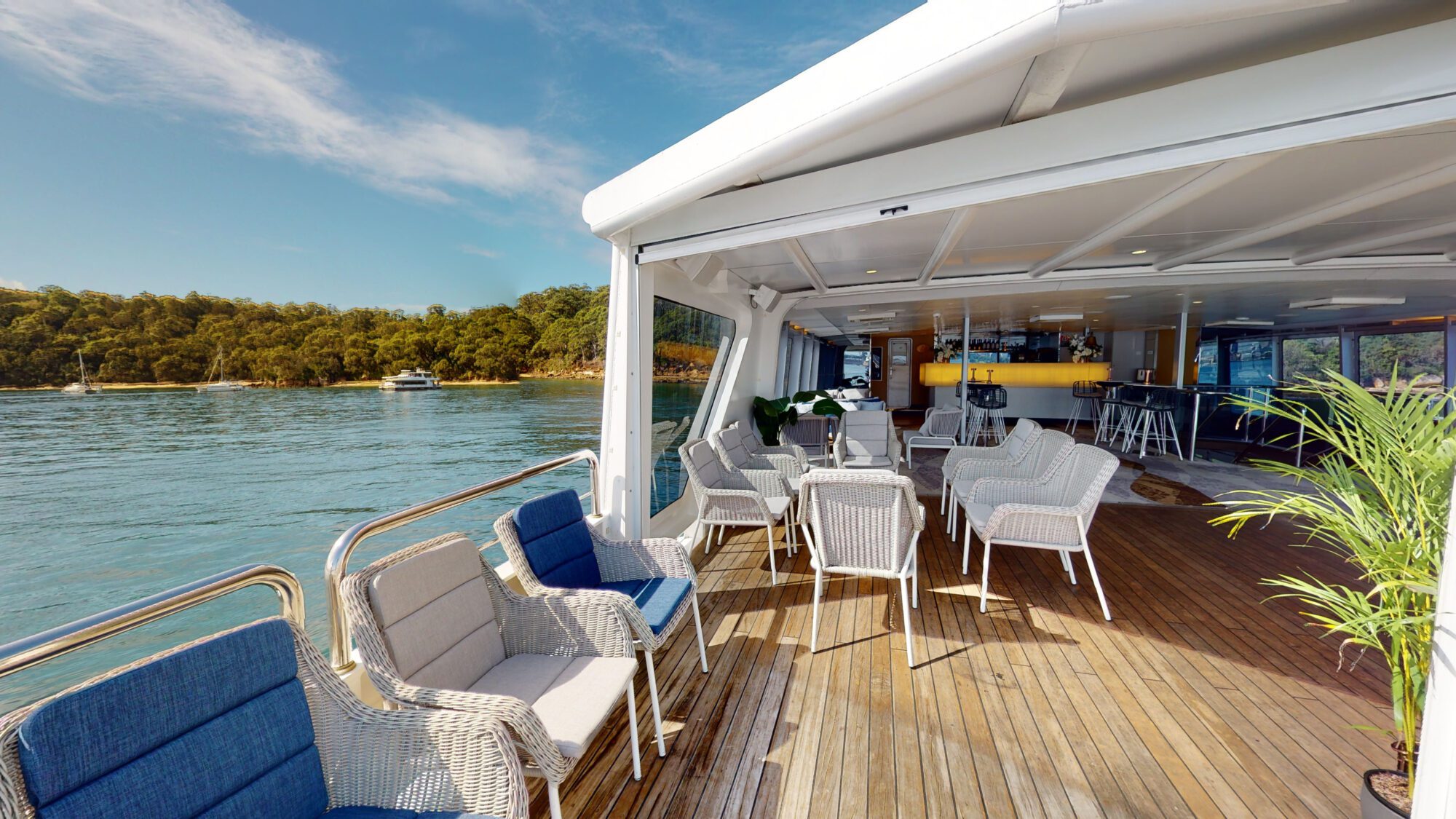 Open-air deck of a boat with wicker chairs and blue cushions, overlooking calm water and a wooded shoreline. The deck leads to a covered area with bar seating and plants, under a bright blue sky.