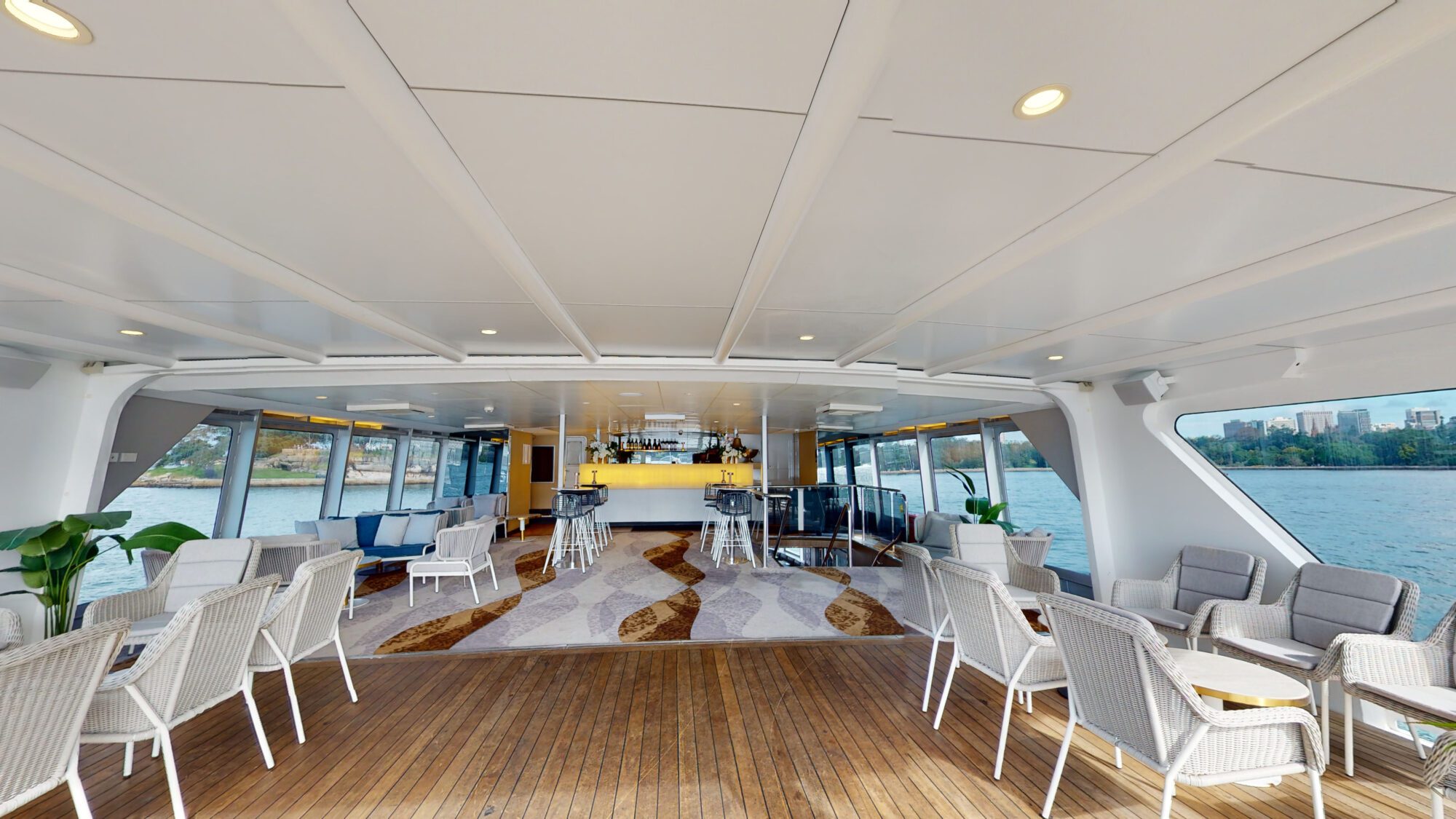Spacious yacht interior with white chairs and tables, wooden flooring, a modern bar at the back, large windows revealing water and city buildings outside, and potted plants as decor.