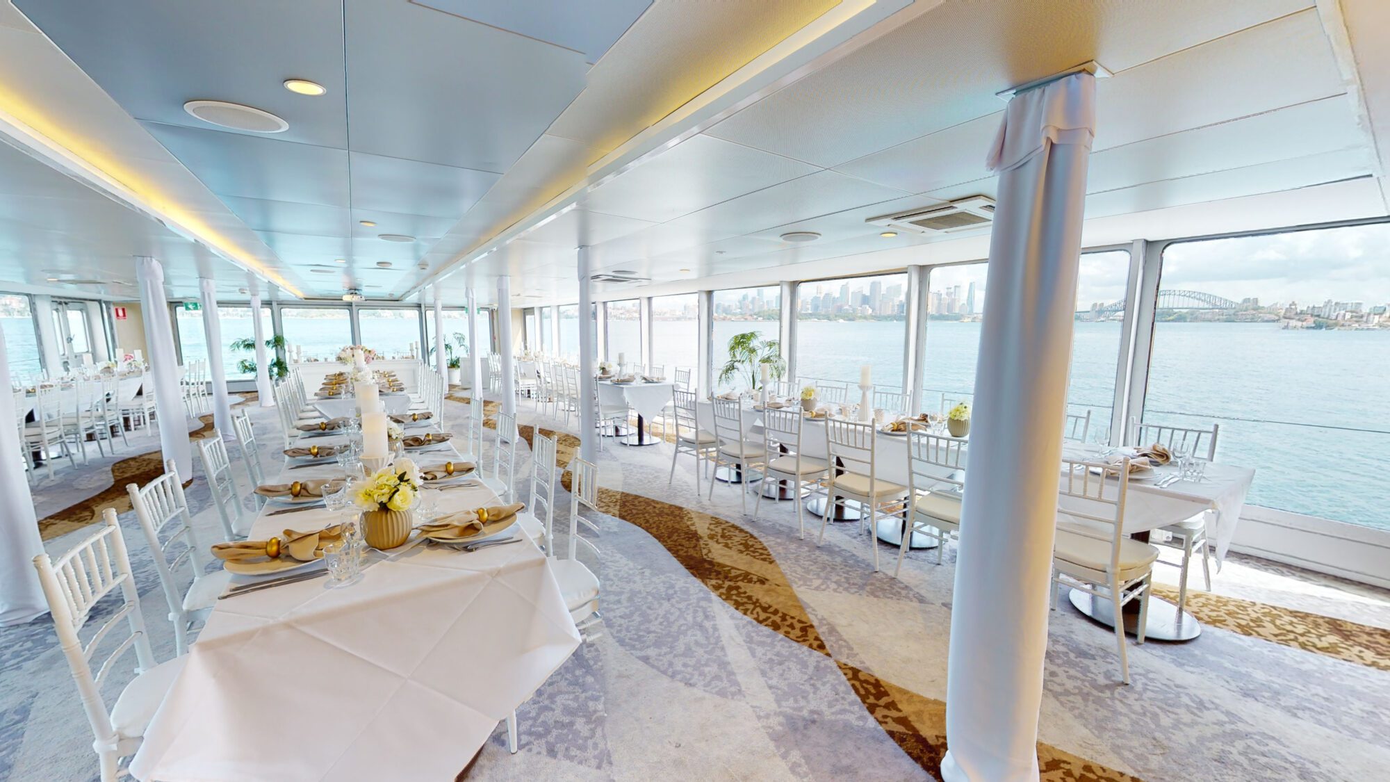 Elegant dining area on a boat with white tables and chairs, floral centerpieces, and large windows offering panoramic views of the water and city skyline in the background. Natural light fills the spacious, modern interior.