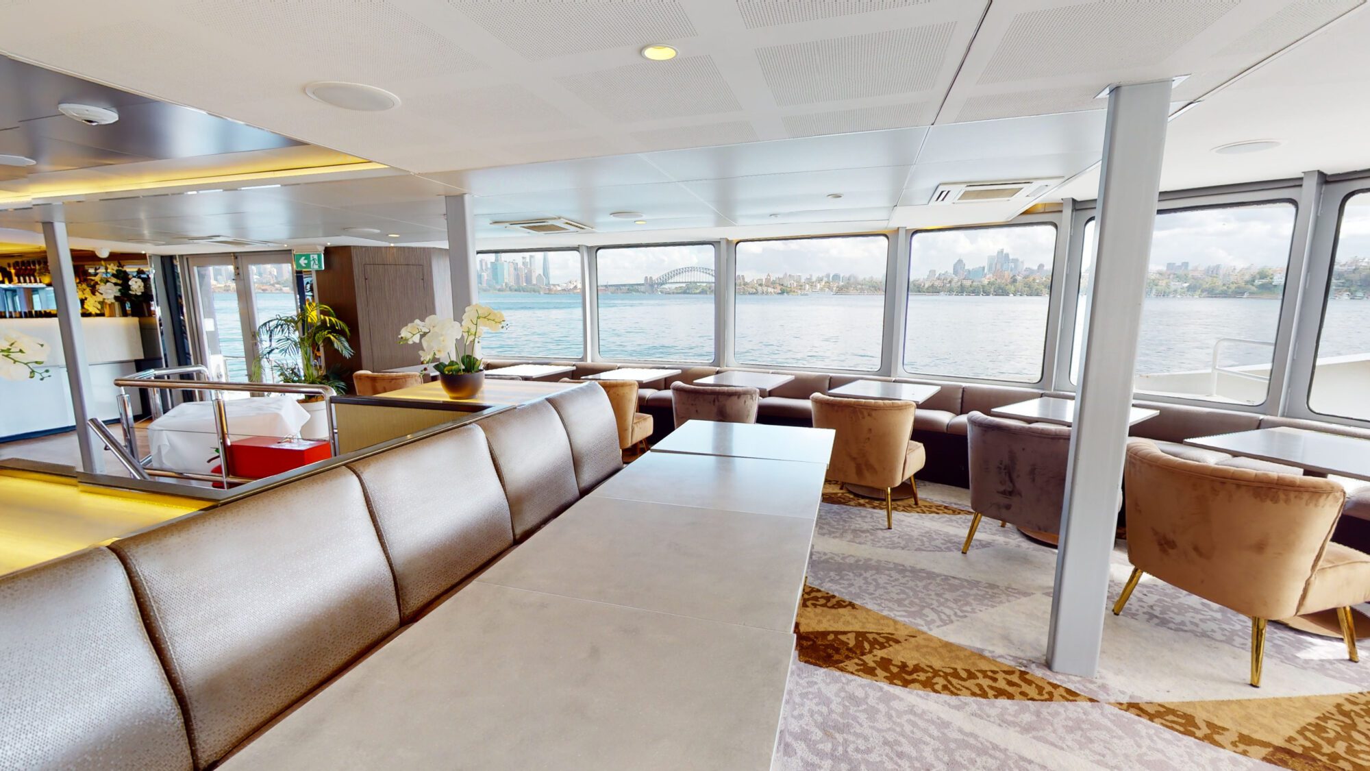 Modern ferry interior with cushioned seats, small tables, and large windows offering views of the city skyline and water outside. Neutral tones and decorative plants create a comfortable, inviting atmosphere.