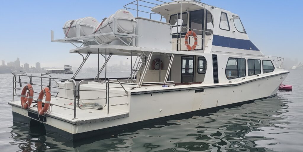 A white motorboat with two decks and several life rings floats on calm water, with a city skyline visible in the distance under a blue sky.