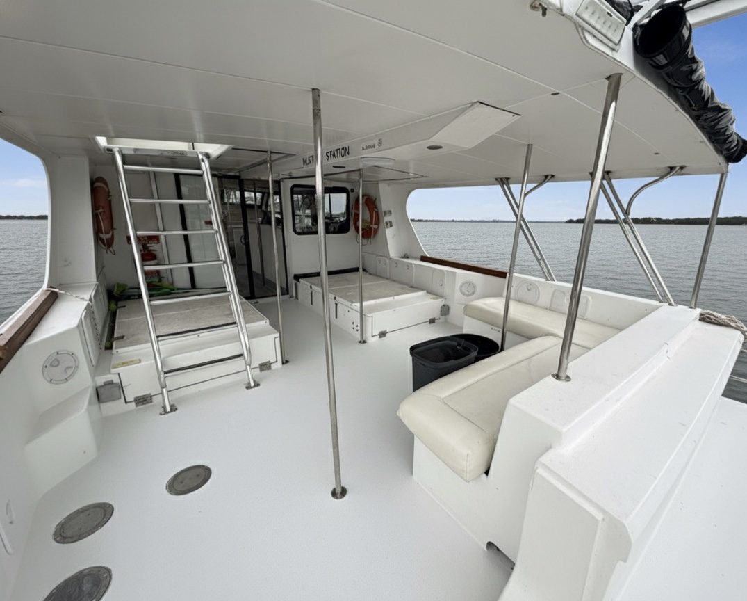 The interior deck of a white boat with cushioned benches, metal poles, a ladder leading to an upper level, windows, and life preservers along the walls, floating on calm water under a cloudy sky.