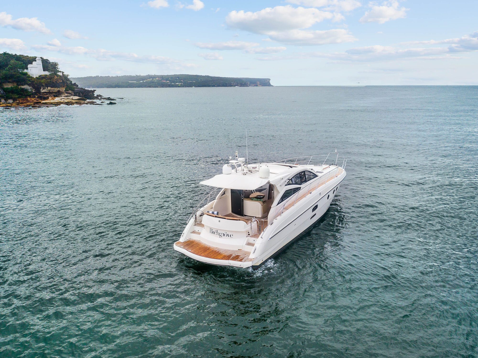 A white luxury motor yacht named "Birchgrove" cruises on calm blue water near a rocky coastline with greenery under a partly cloudy sky.