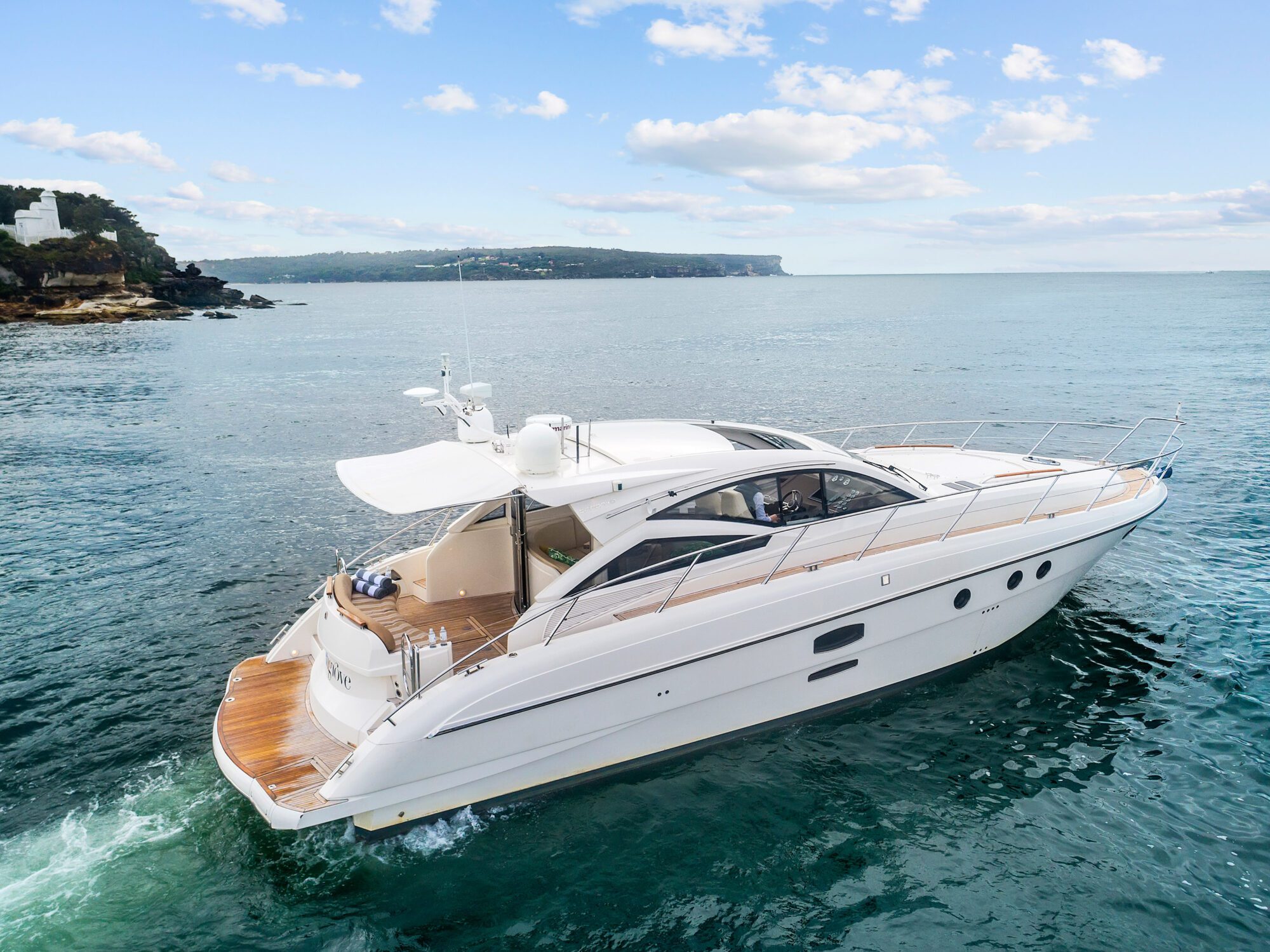 The Birchgrove, a sleek white yacht, glides across calm blue water near a rocky coastline under a partly cloudy sky, distant land visible.
