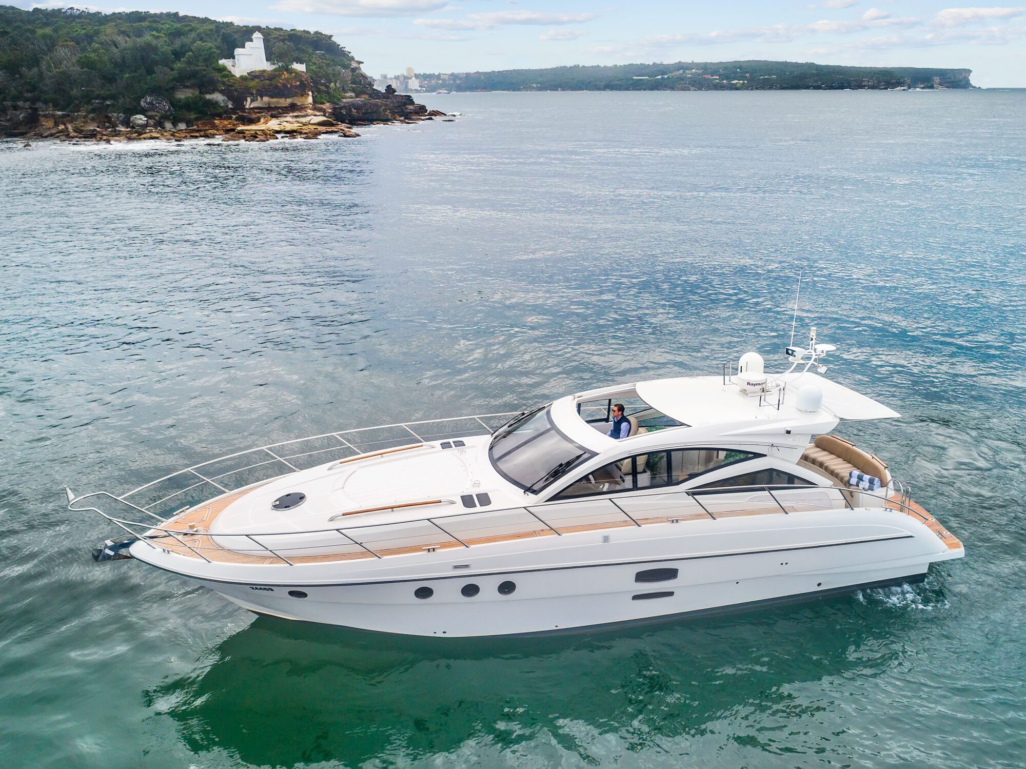The Birchgrove, a sleek white yacht, glides on calm blue-green water near a rocky shoreline with trees and buildings in the background under a partly cloudy sky.