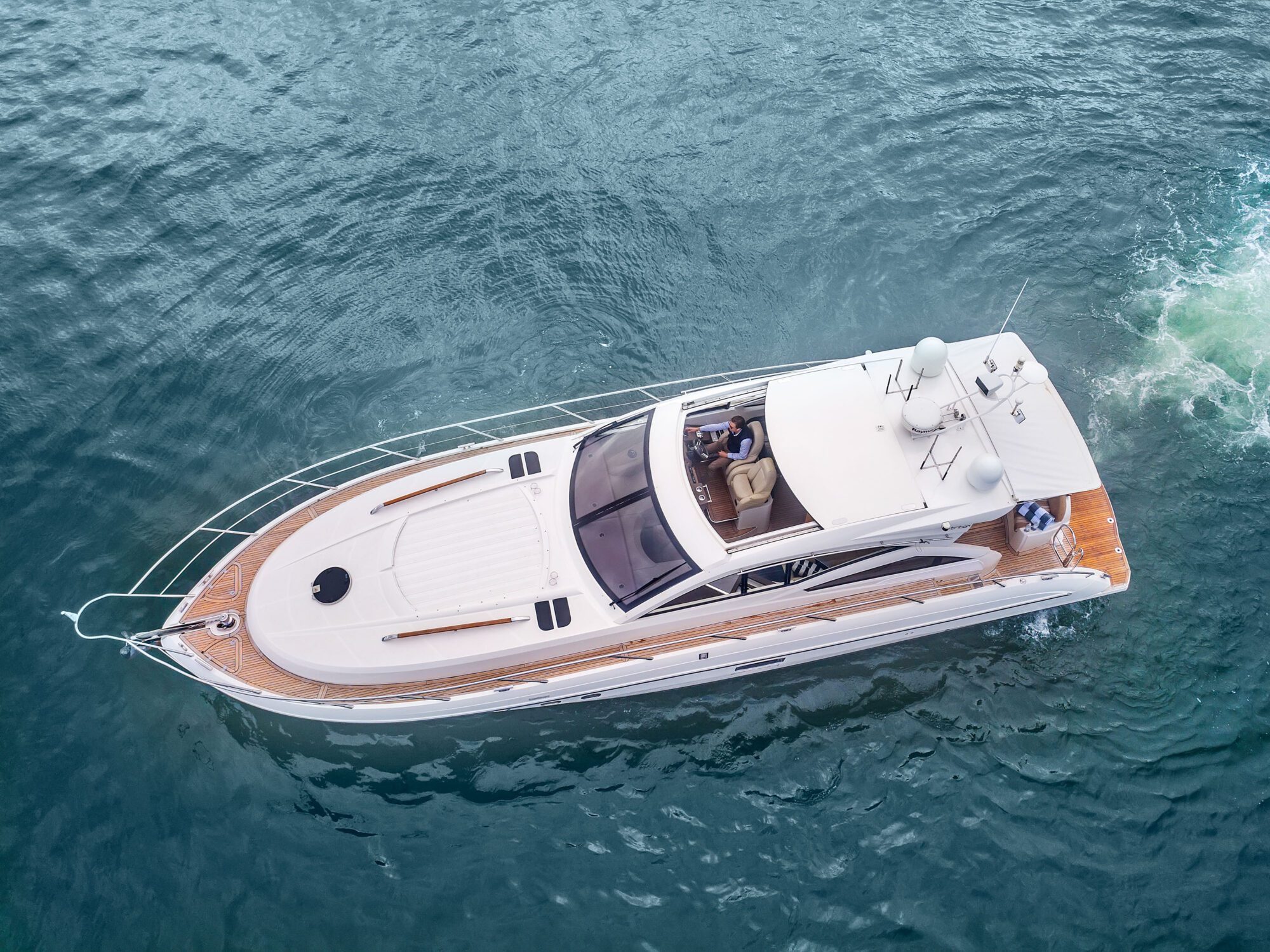 The Birchgrove, a white luxury yacht, cruises through clear blue water as seen from above. Two people are seated in the cockpit while the yacht leaves a trail behind it.