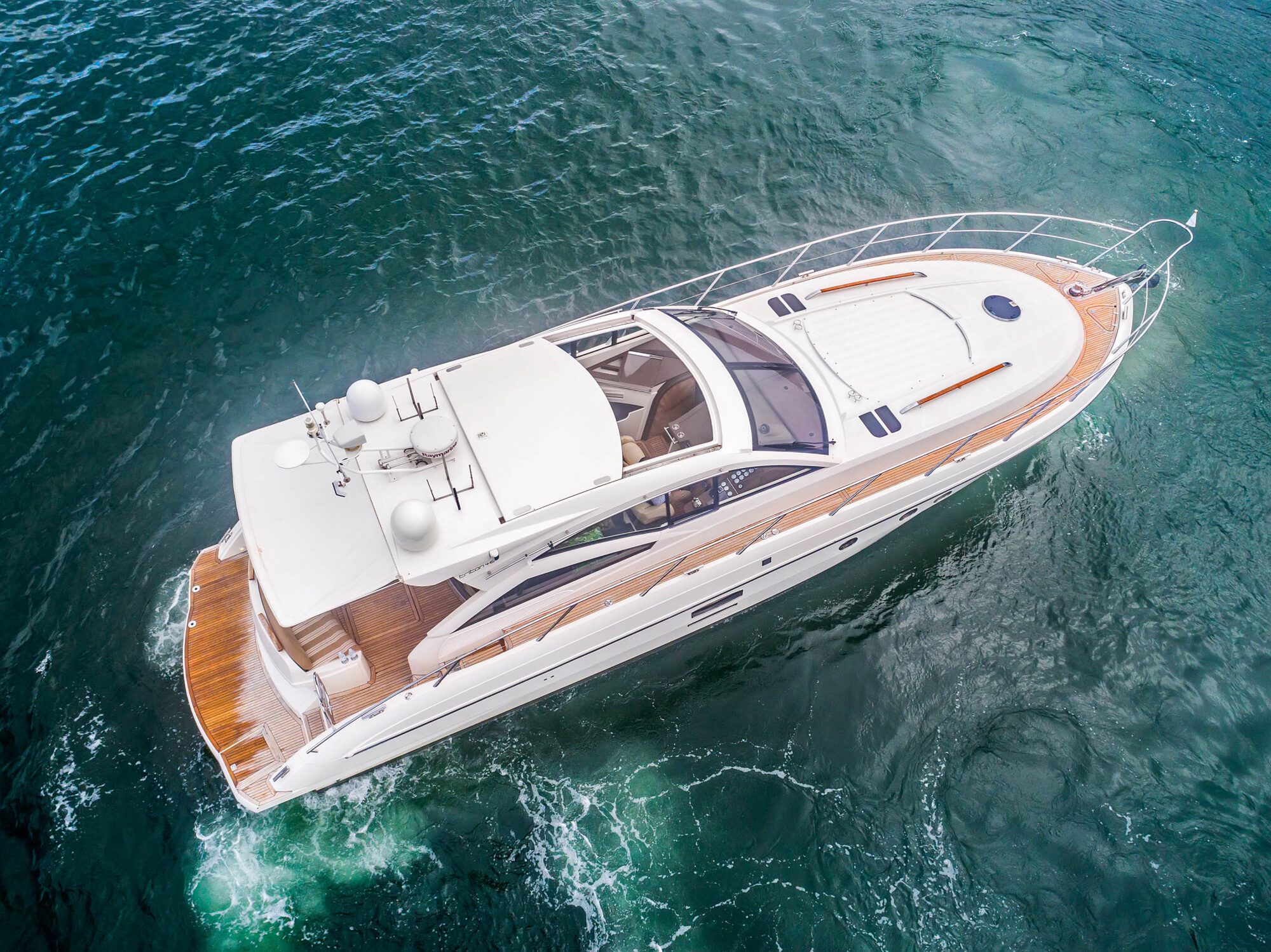 Aerial view of Birchgrove, a white luxury yacht with a wooden deck and sleek, modern design, cruising on clear teal water and leaving a trail of white foam behind.