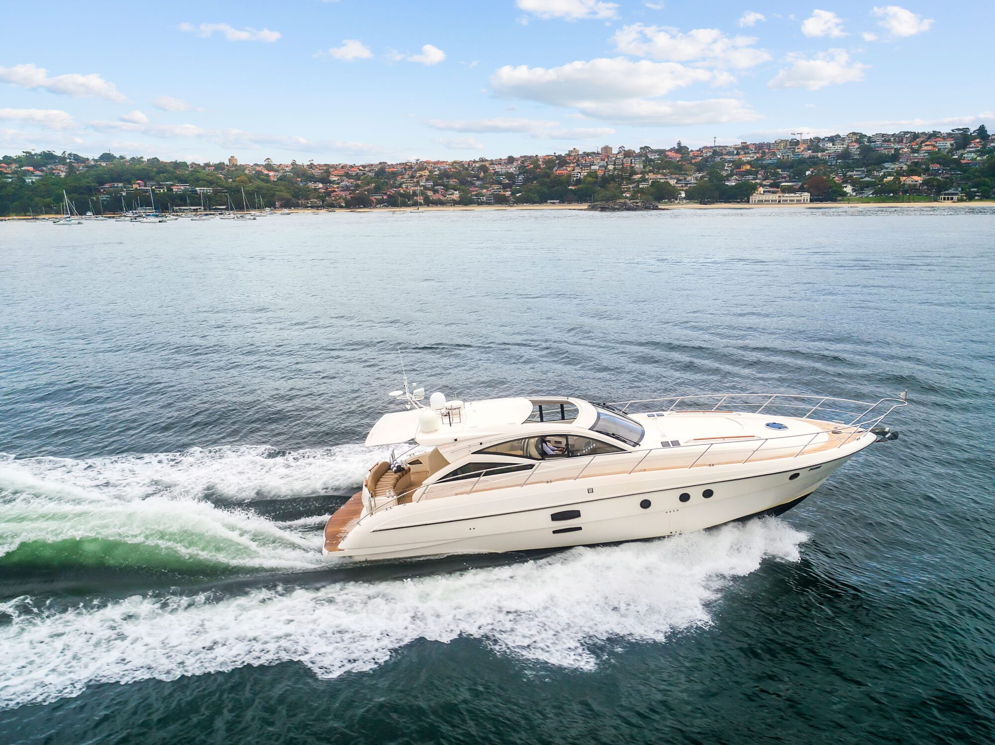 The Birchgrove, a white motor yacht, speeds through calm waters near the shore, leaving a wake. In the background are trees, houses, and a partly cloudy sky.
