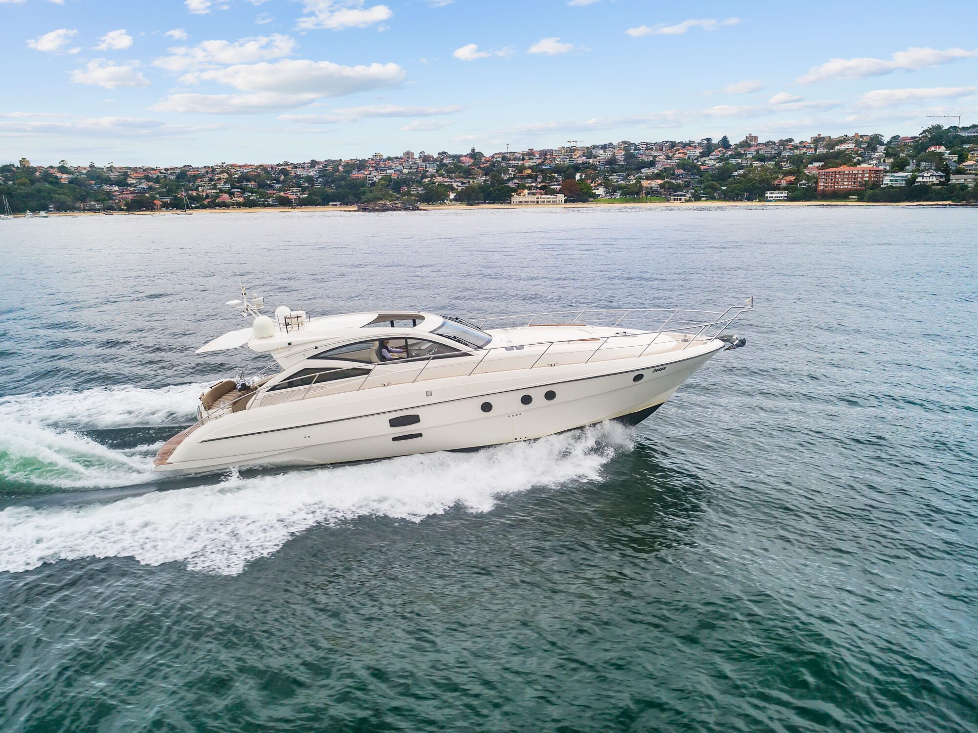 The Birchgrove, a white luxury yacht, speeds across calm blue water near a coastline lined with buildings and trees beneath a partly cloudy sky.