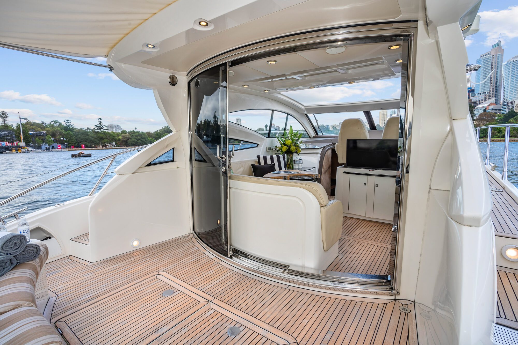 Experience Birchgrove’s open deck with teak flooring, cushioned seats, and glass doors revealing an elegant interior lounge with a dining table, fresh flowers, and a cityscape view over calm water.
