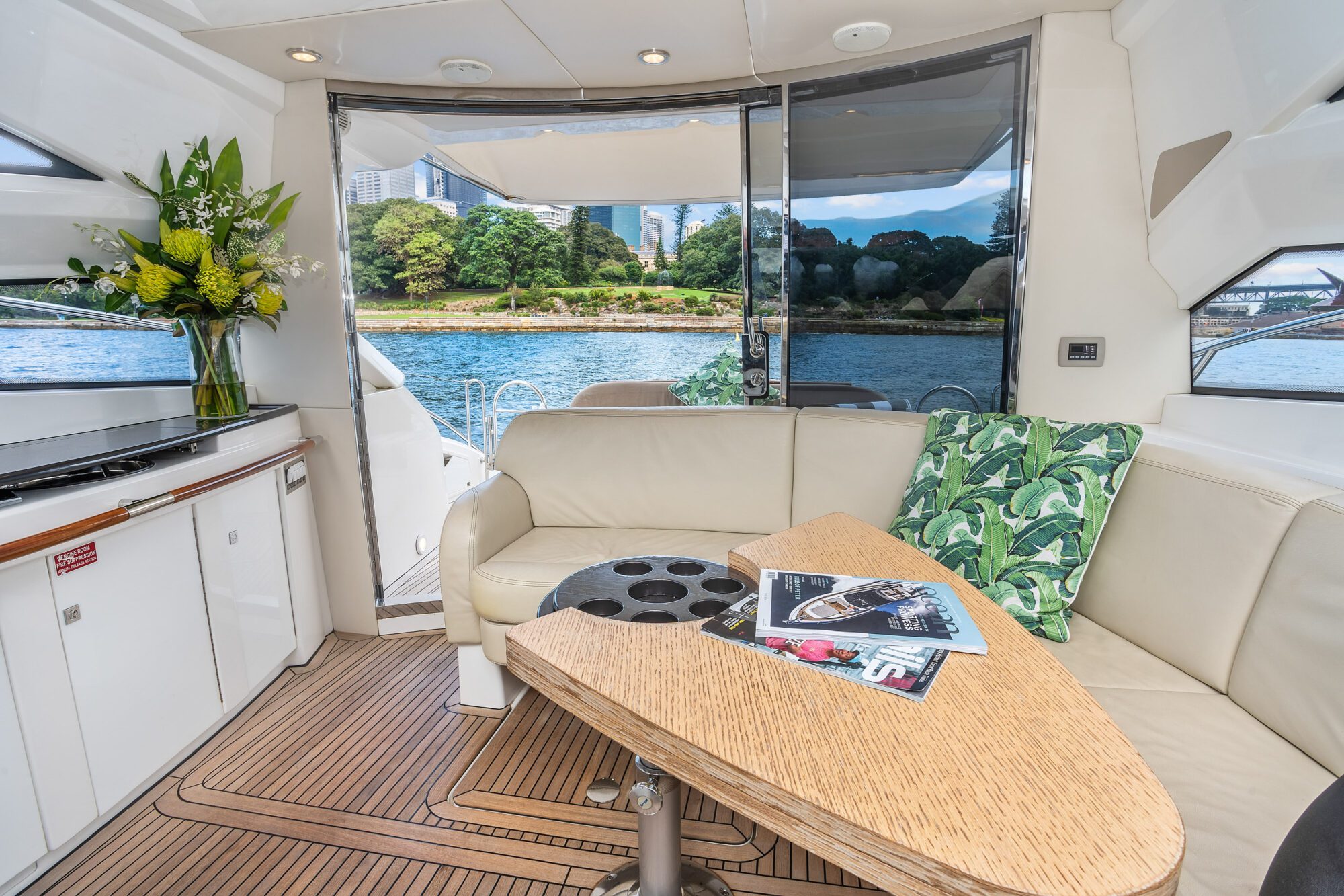 Birchgrove yacht interior with cream seating, a wooden table with magazines, and green leaf-print pillows. Large windows reveal water and trees outside; a bouquet of flowers rests on the left-side cabinet.