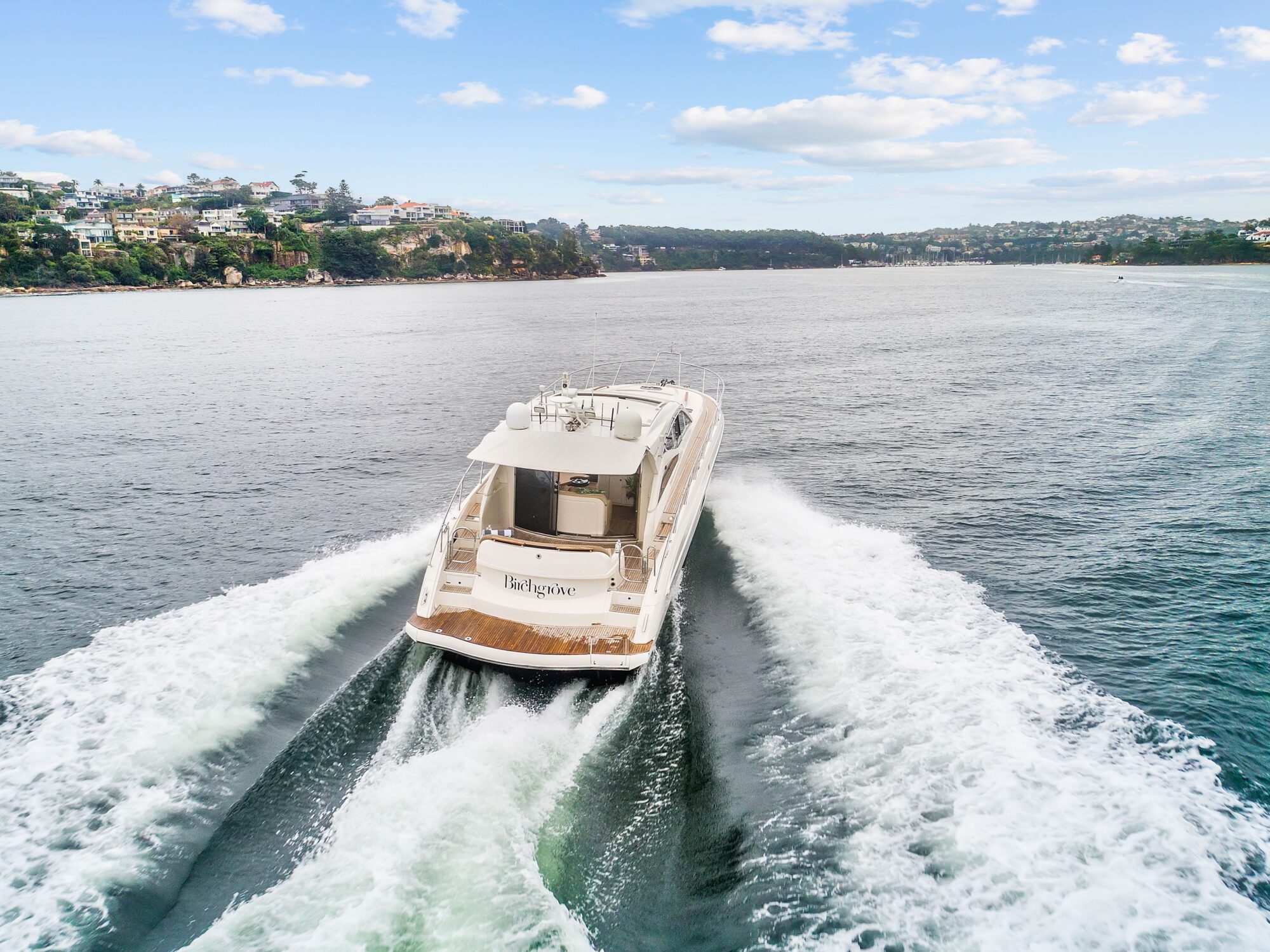 The Birchgrove, a luxury white yacht, glides through calm waters with houses and lush greenery on either side beneath a partly cloudy blue sky.