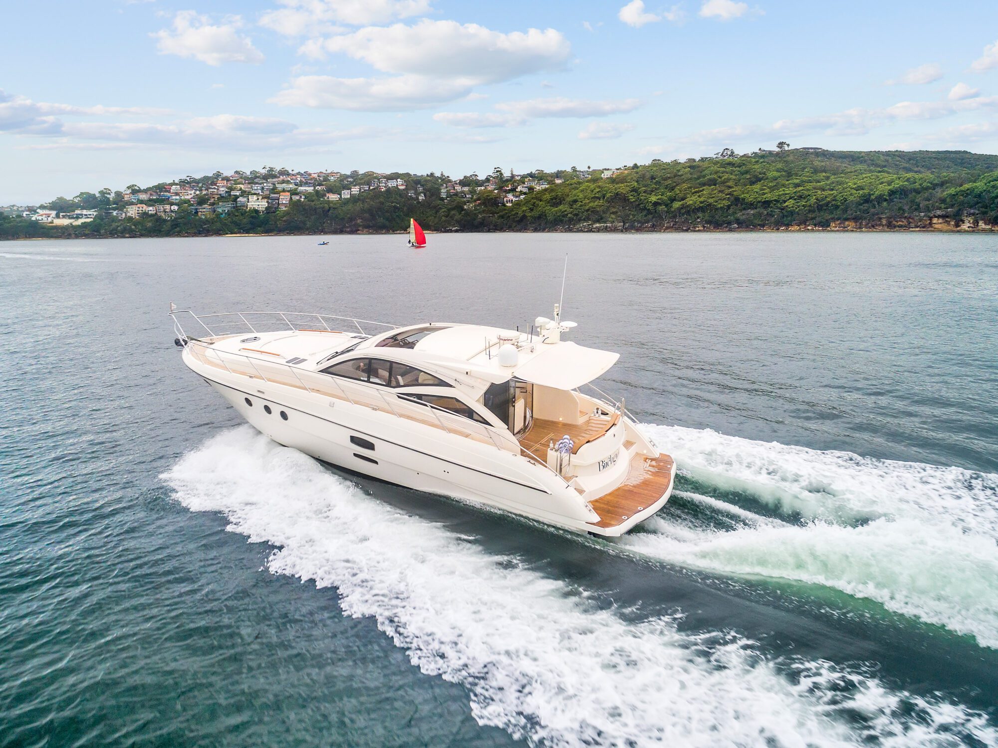 Birchgrove, a white luxury yacht, speeds through calm waters near a green, hilly coast under partly cloudy skies, leaving a wake behind; houses dot the distant hillside.