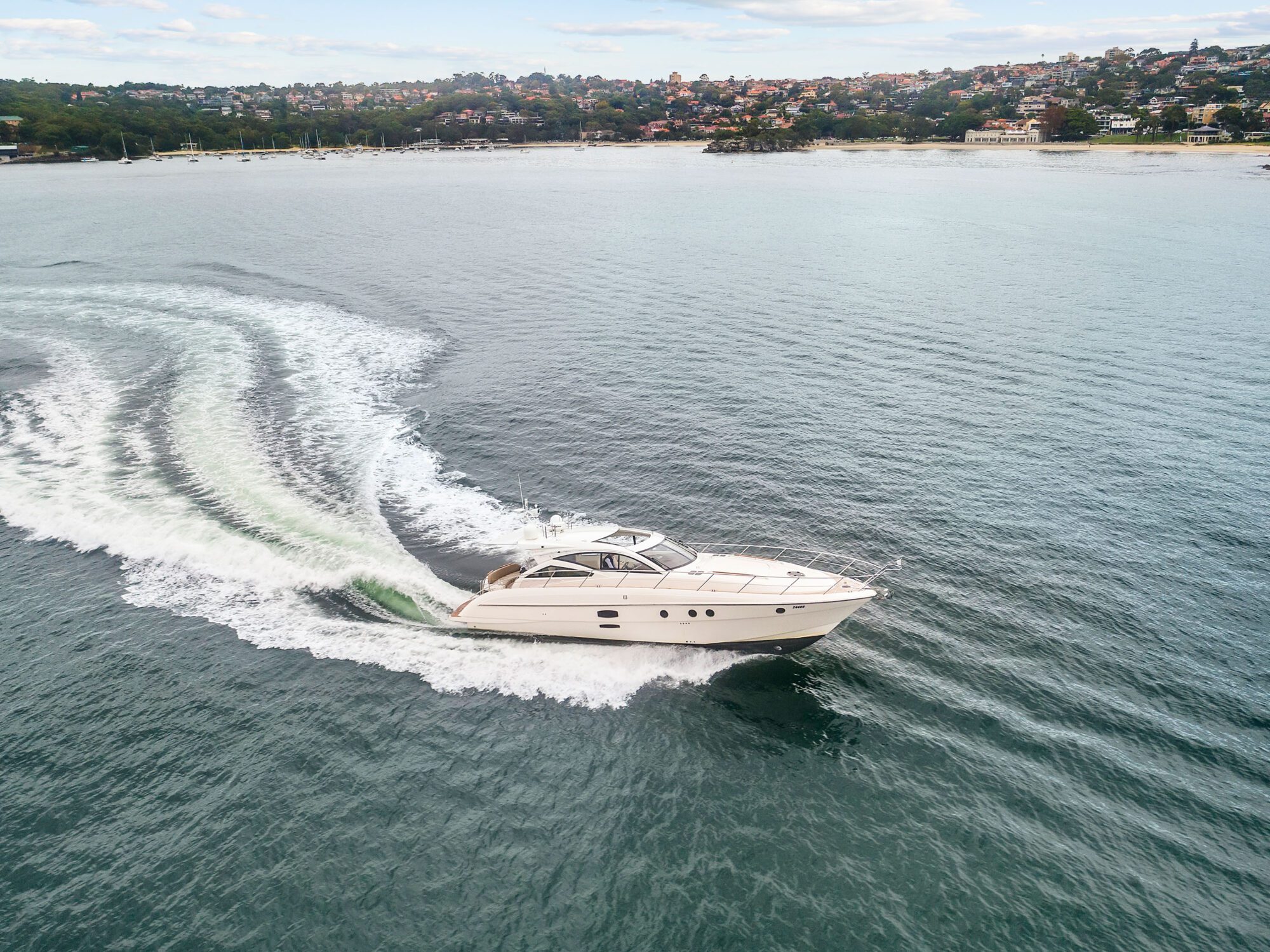 The Birchgrove, a white motor yacht, speeds across calm water leaving a curved wake. In the background, houses and trees line the shore beneath a partly cloudy sky.