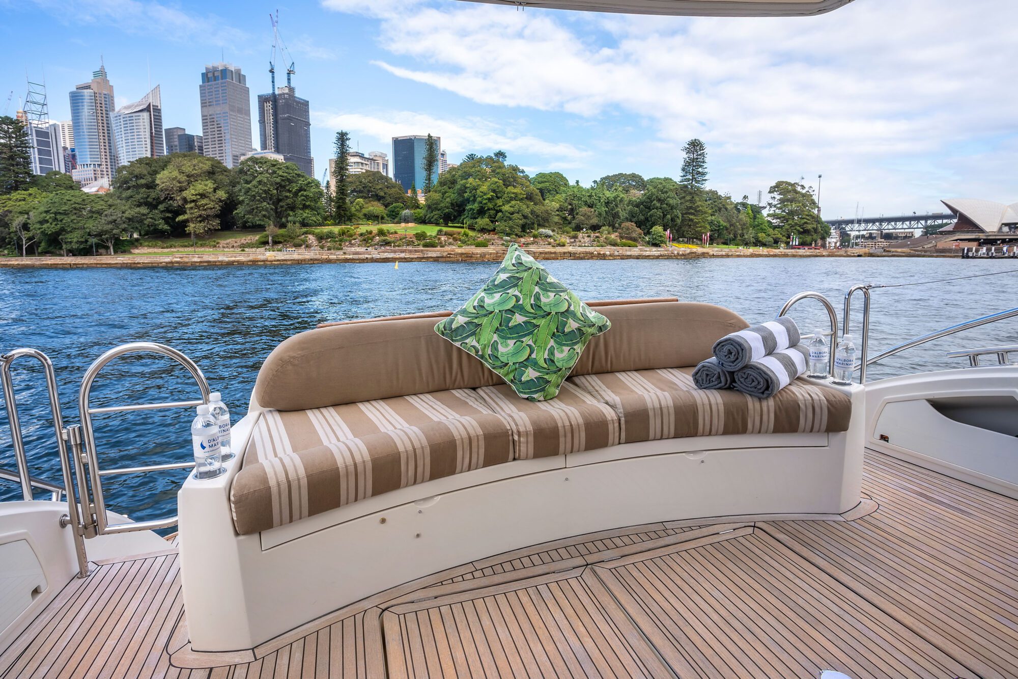 The Birchgrove bench features curved stripes and a green cushion, placed on a boat deck with rolled towels, overlooking blue water and a city skyline with trees and tall buildings in the background.