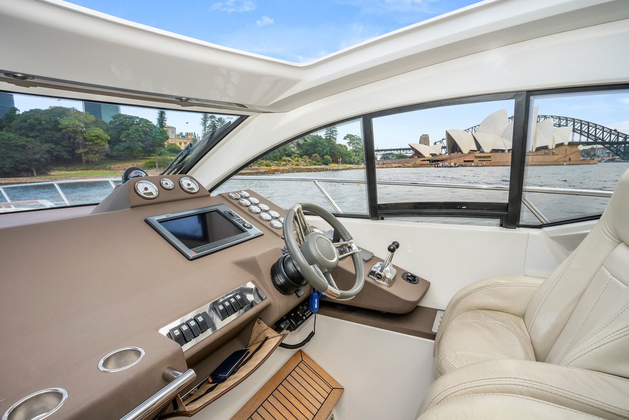 The interior of the Birchgrove yacht features a helm with leather seats and navigation controls; through the windows, the Sydney Opera House and Harbour Bridge appear against a blue sky.